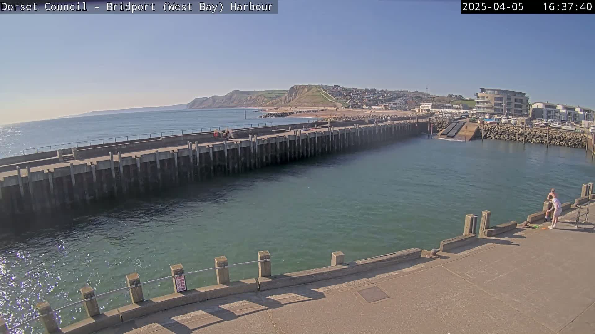A harbor with a long concrete pier juts into calm, greenish-blue water under a clear, sunny sky, with a town and cliffs visible in the distance.