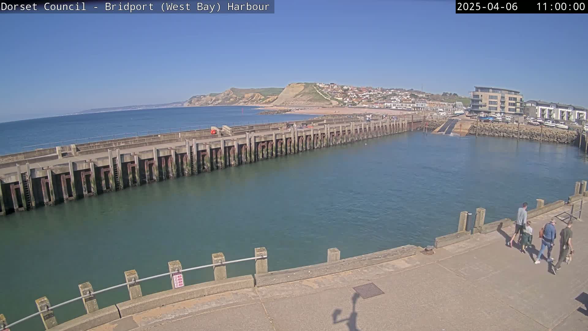 A harbor with a long concrete pier extending into calm, blue water under a clear, sunny sky, with a small group of people walking along a paved area near the water's edge and a town visible in the background.