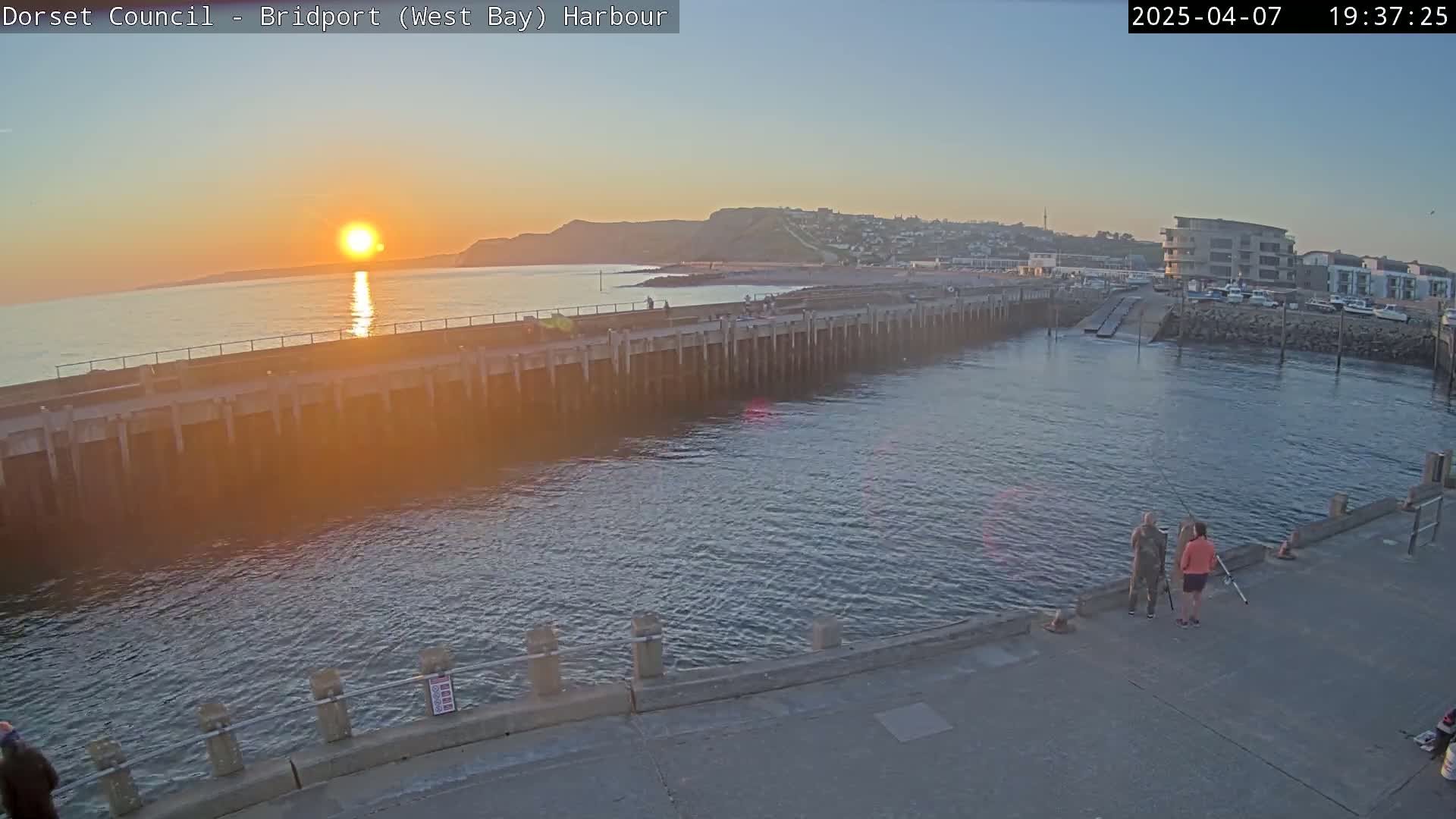 A harbor at sunset, with a long pier extending into calm water, shows two people fishing on a pier in the foreground.