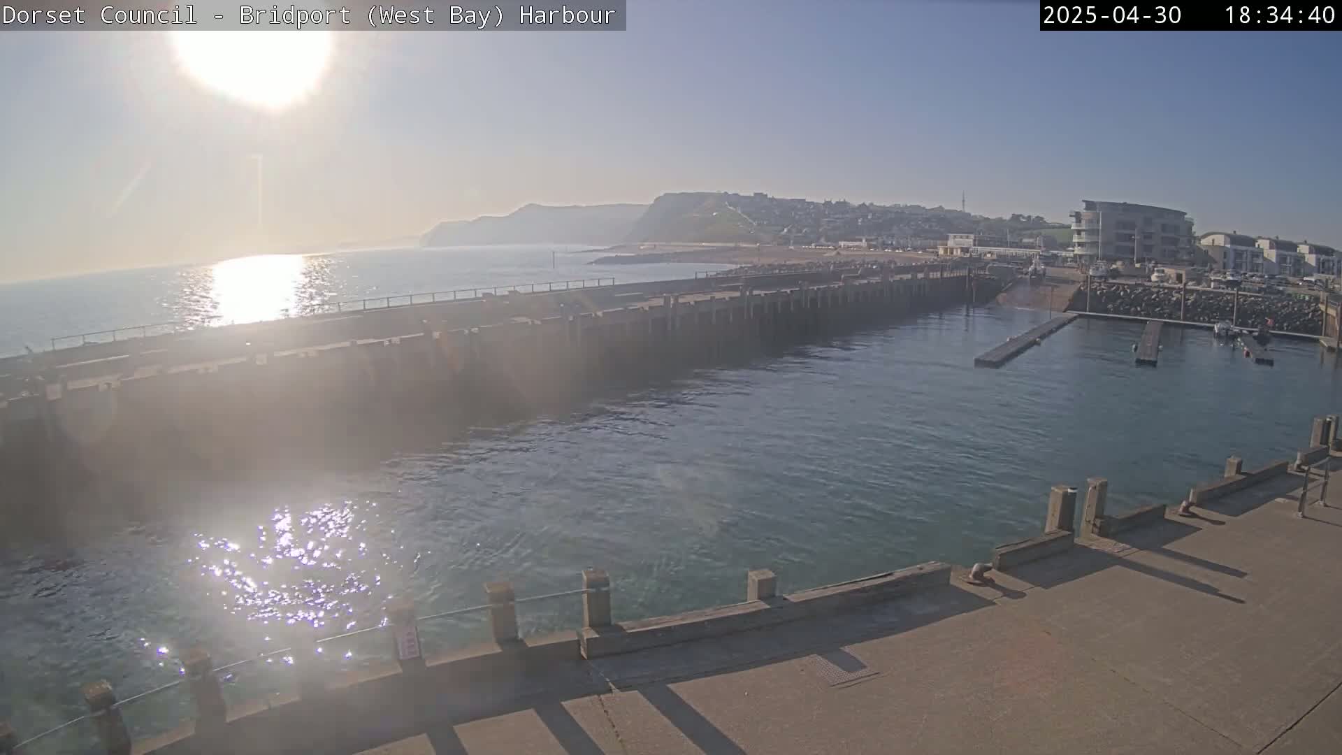 A harbor with calm water reflecting sunlight, a breakwater, and buildings in the background under a clear, sunny sky.