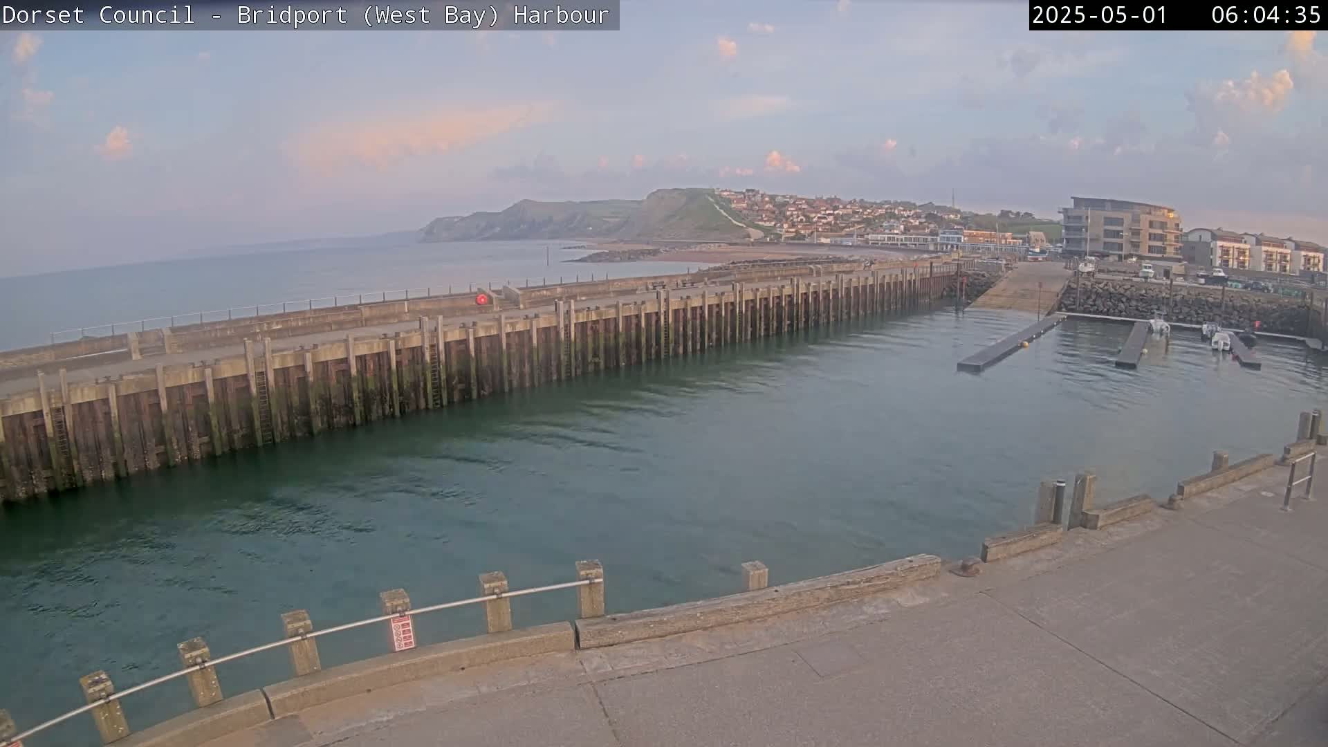 A harbor with calm water under a partly cloudy sky shows a long pier extending into the water, with a small town and hills visible in the distance.