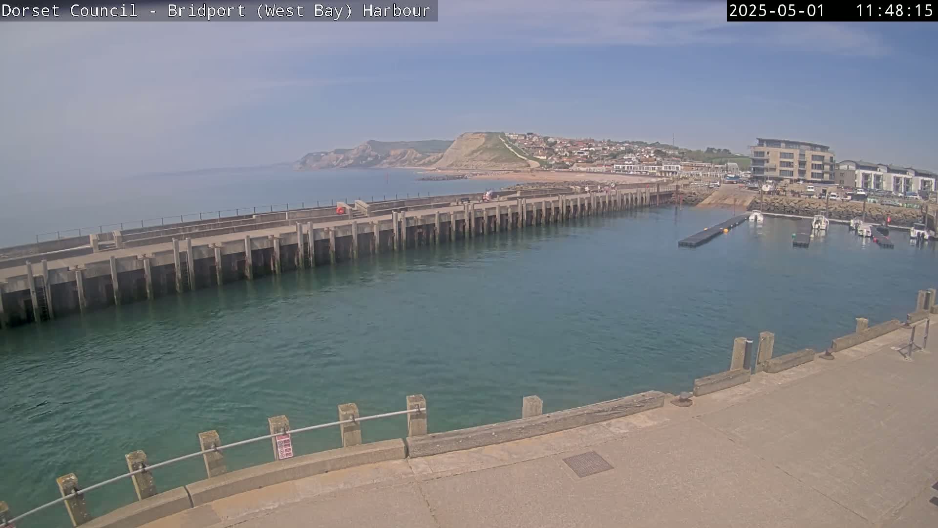 A harbor with a long concrete pier extends into calm, teal water, under a sunny sky, with a town and cliffs visible in the distance.
