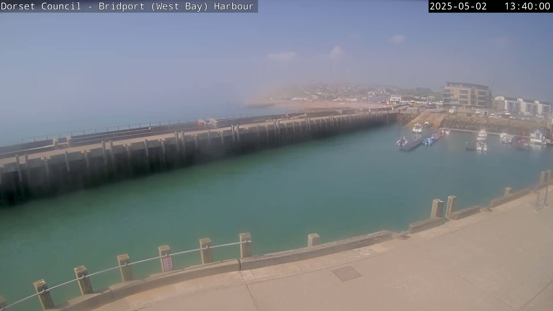 A hazy, sunny day reveals a harbor with several small boats moored, nestled between a long, concrete wall and a shoreline with buildings visible in the distance.