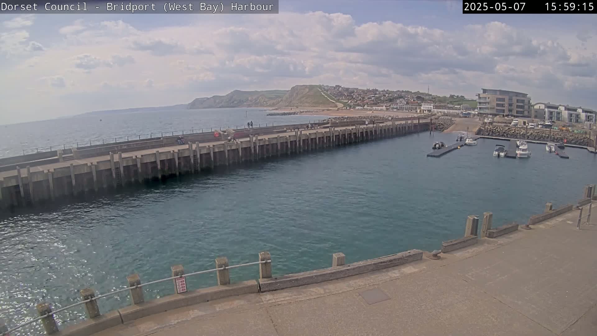 A harbor with several small boats moored, a long concrete pier, a town on a hillside in the distance, and a partly cloudy sky.
