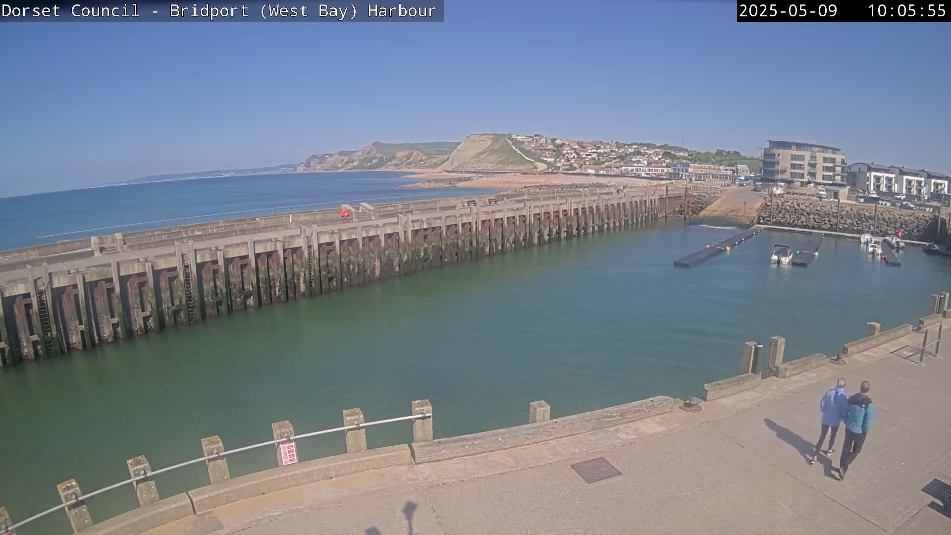 A harbor with a long concrete pier, a calm body of water, a few boats, and a sandy beach with cliffs in the background on a sunny day.