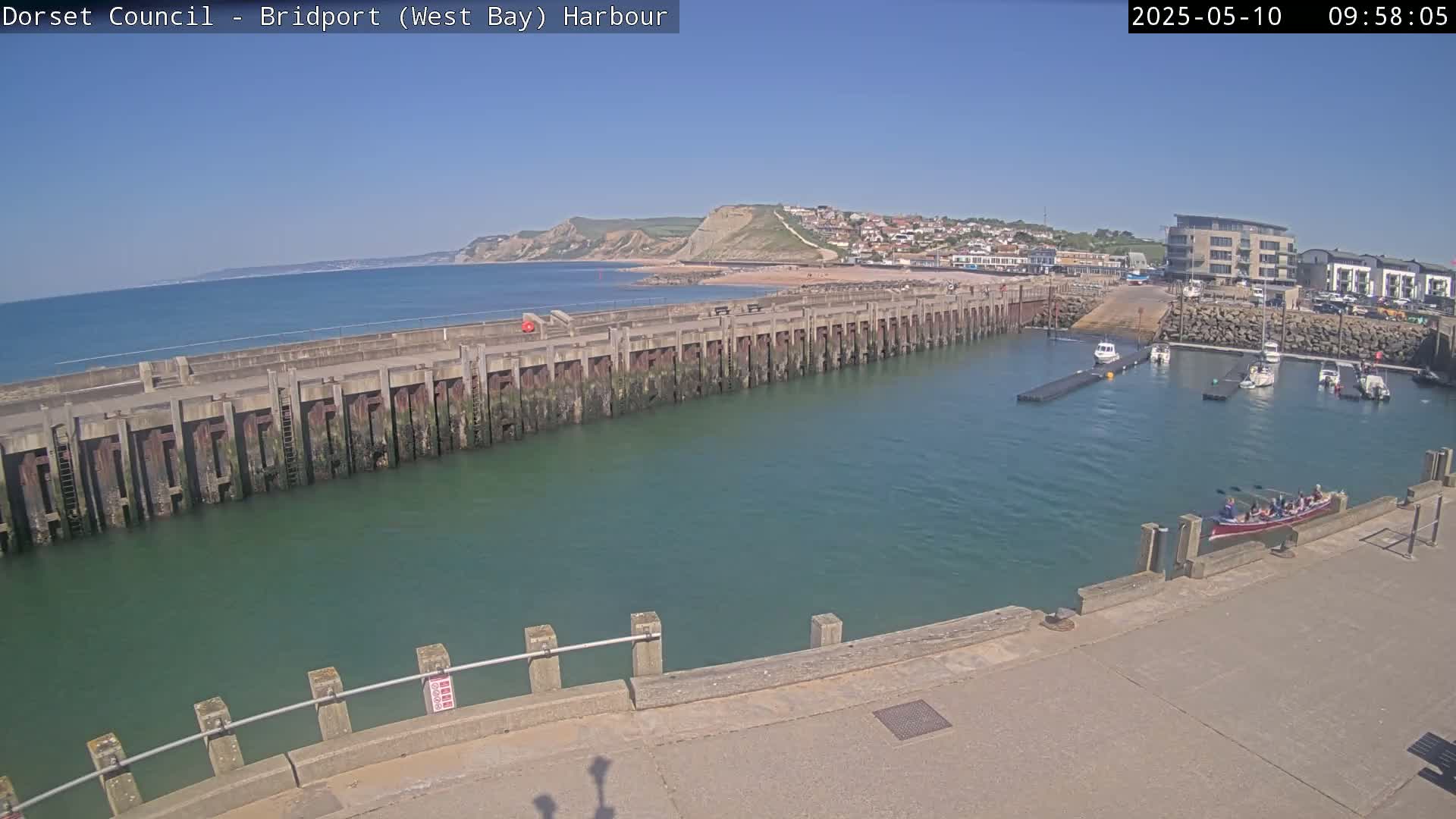 A harbor with a long pier, several boats, and a small town nestled against a cliffside on a sunny day.