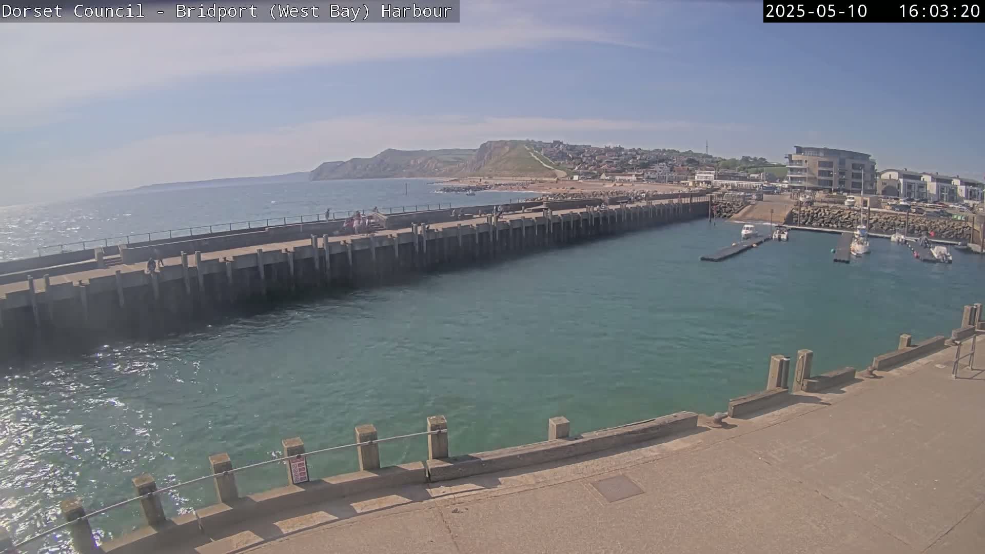 A harbor with a long concrete pier, a calm body of water, several boats docked, and a hilly coastline visible in the background under a mostly sunny sky.