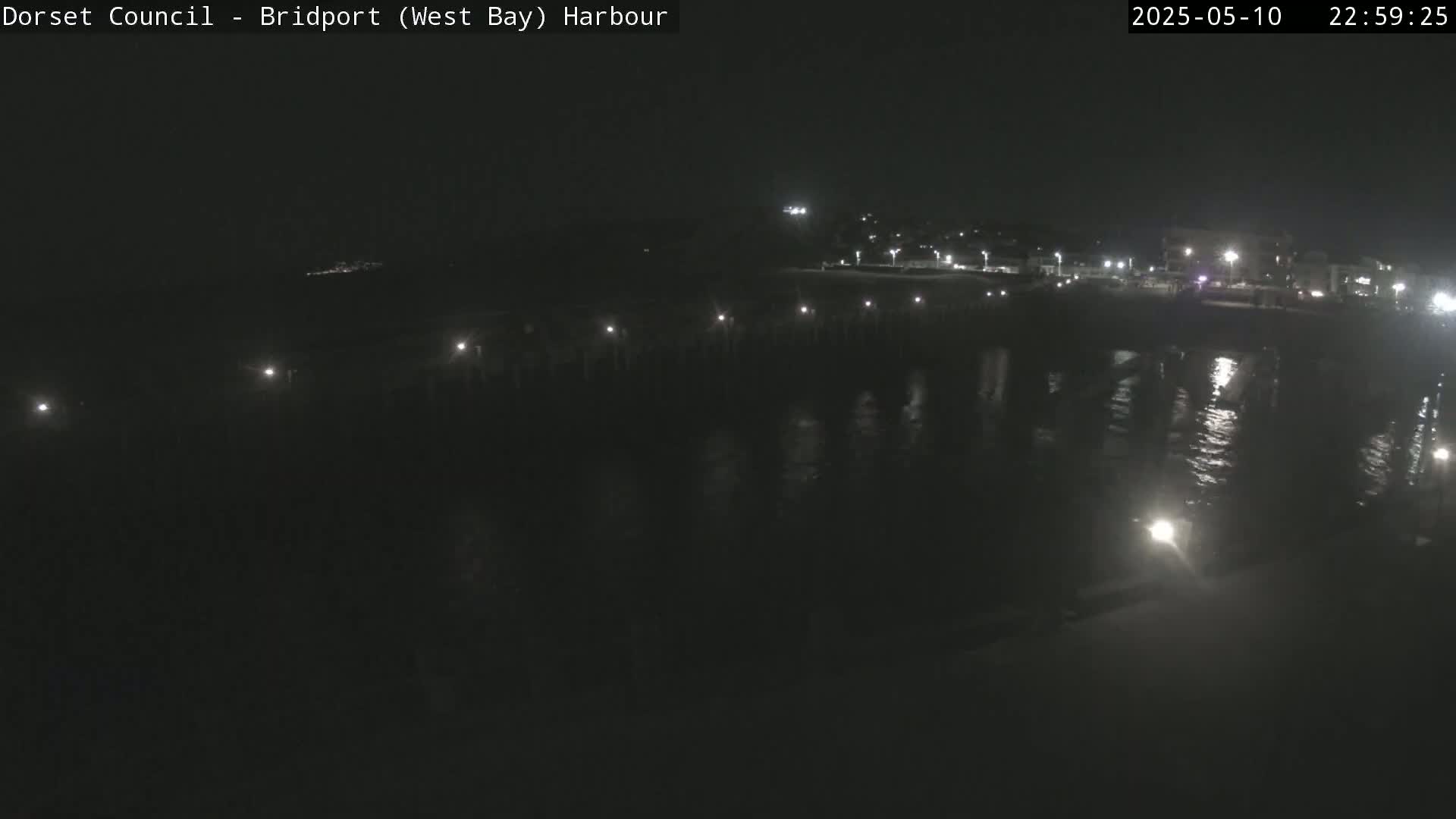 A nighttime harbor scene shows a long pier extending over dark water reflecting lights from the pier and a distant shoreline.