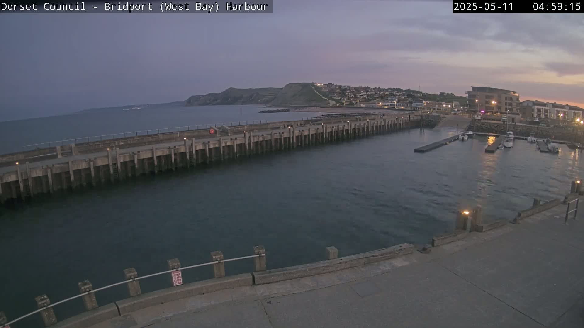 A harbor at dawn shows a calm body of water, a long concrete pier, and several boats docked in a marina, with a hill and town visible in the background under a dusky sky.