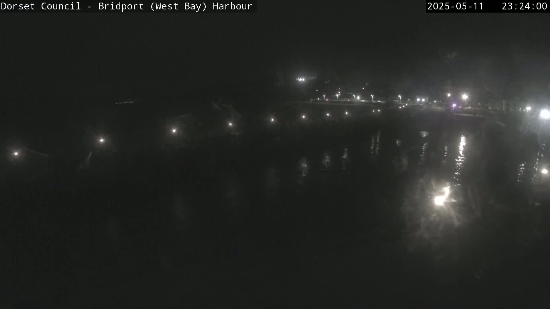 A dark nighttime harbor scene shows a calm body of water reflecting distant lights from a shoreline.