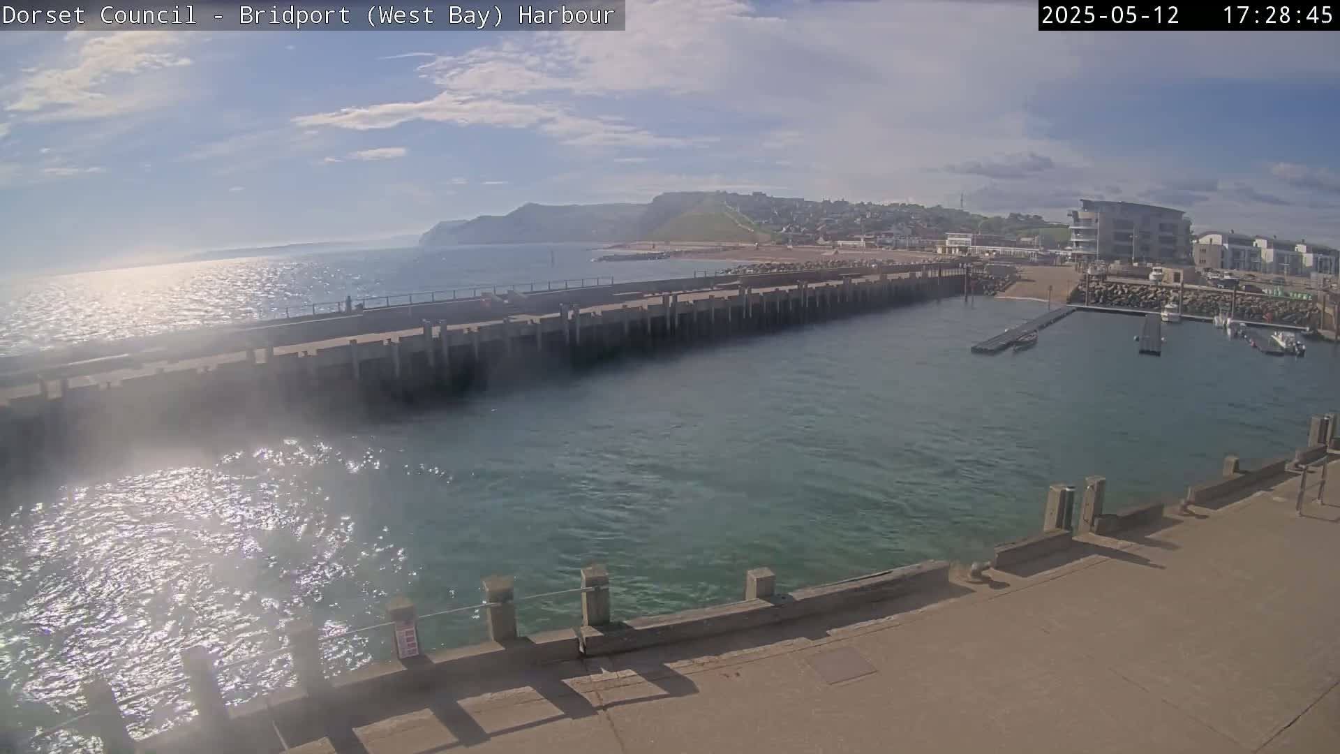 A harbor with a long concrete pier extends into a calm, sunlit sea, backed by a coastline with buildings and hills under a partly cloudy sky.