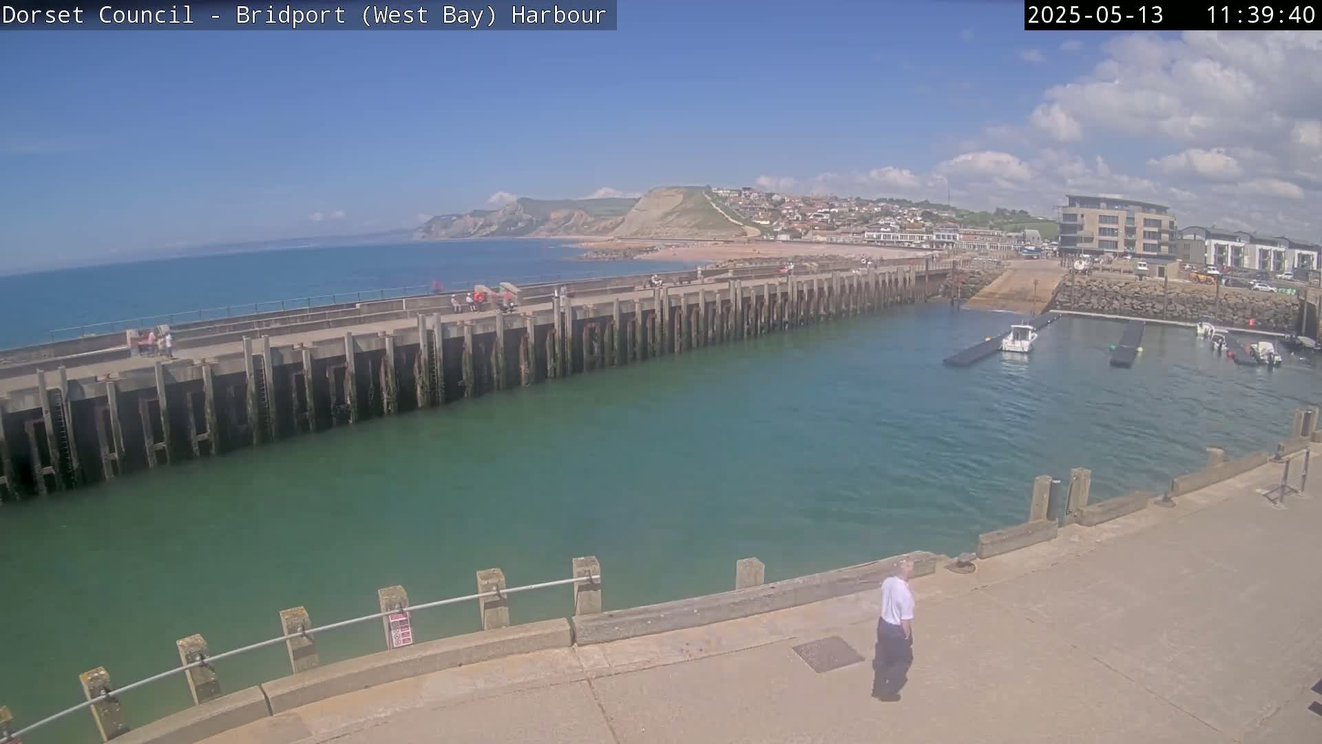 A harbor with a long pier, boats in the water, a person walking on a paved area, and a beach and cliffs visible in the distance under a mostly sunny sky.
