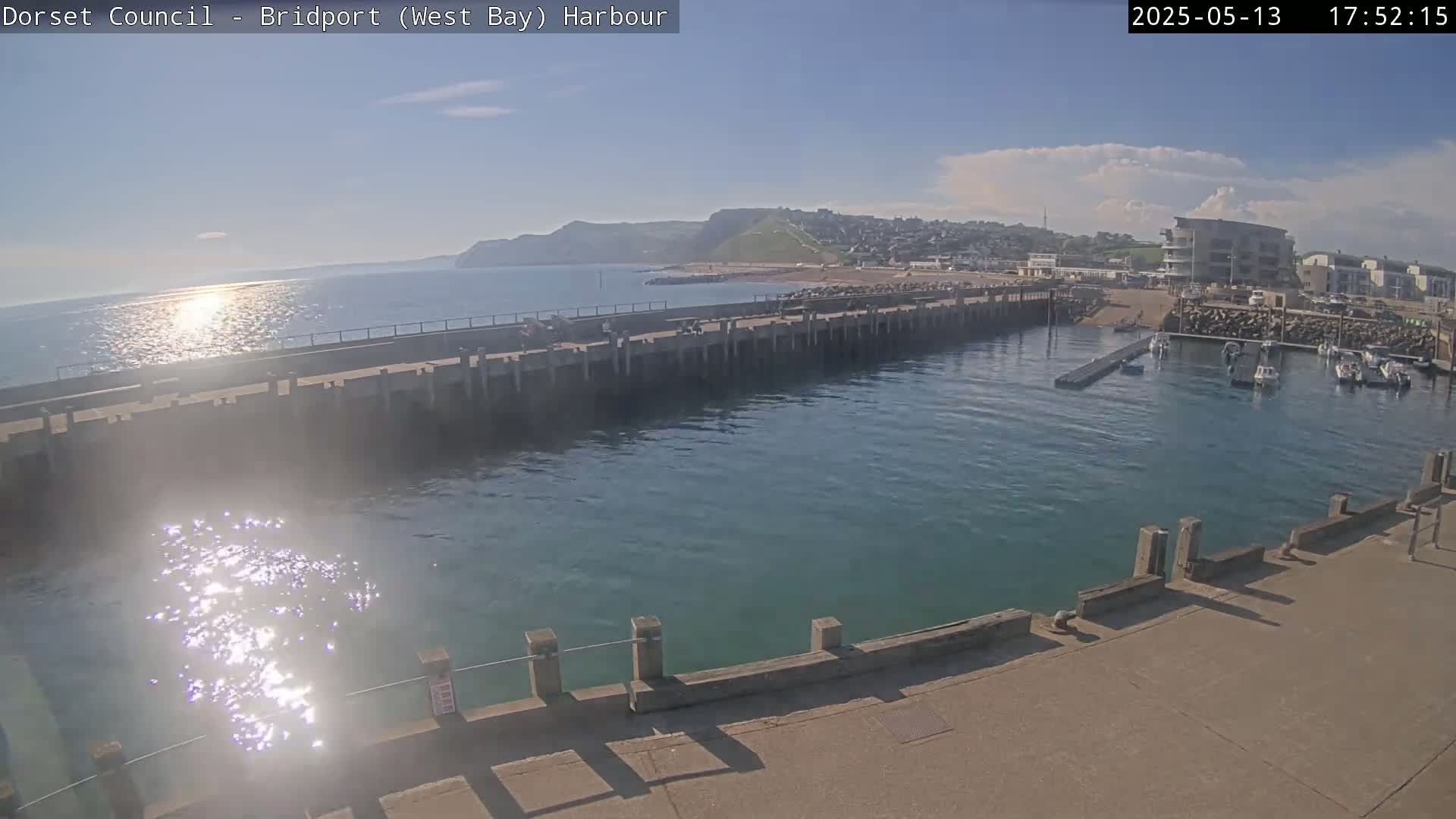 A harbor with calm, sunlit water is partially enclosed by a concrete pier, with boats moored in the harbor and a hillside town visible in the background under a mostly clear sky.