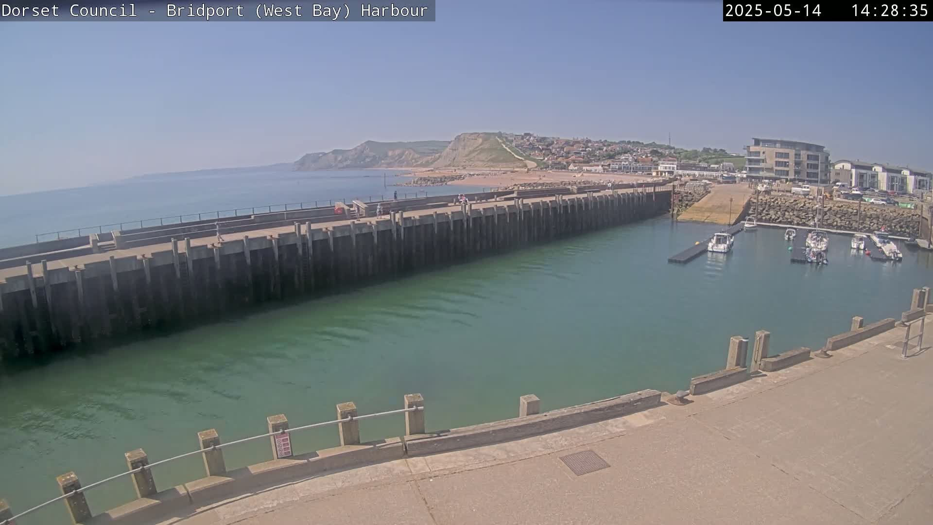 A harbor with several boats is visible, next to a long concrete wall and a sandy beach, all under a clear sunny sky.