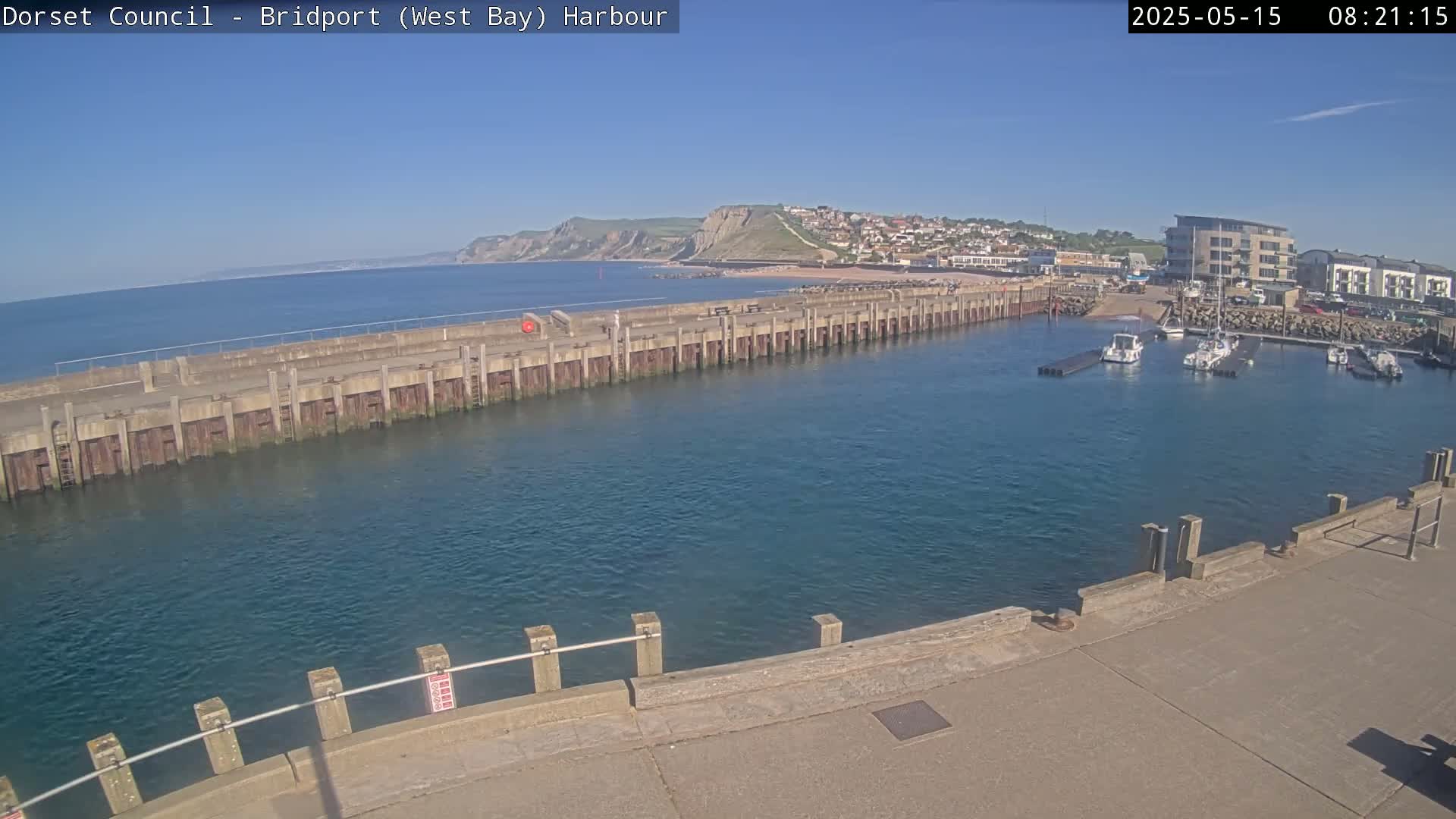 A harbor with calm, blue water is nestled between a long concrete pier and a town backed by cliffs under a clear, sunny sky.