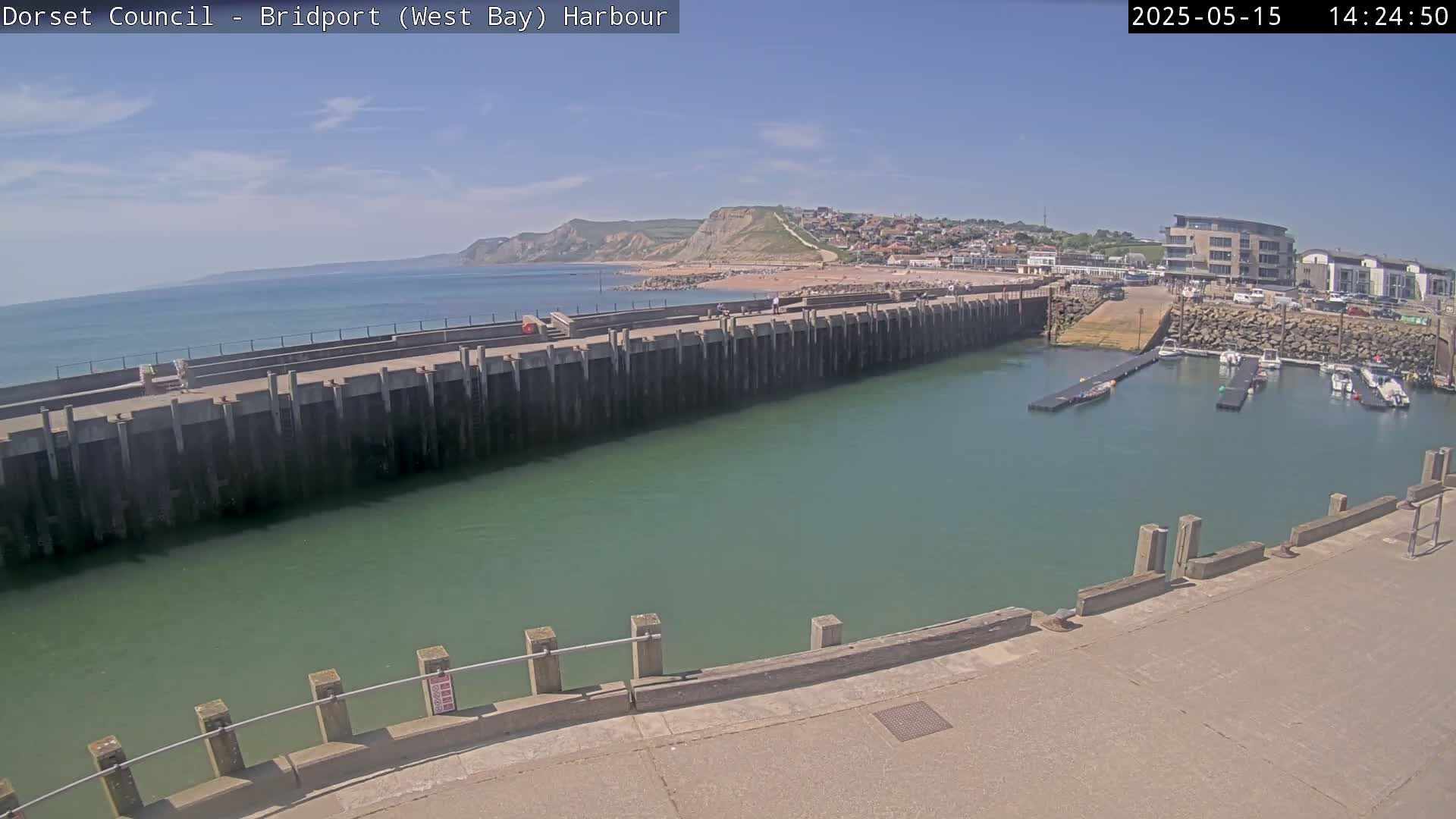 A harbor with a long concrete wall separating the calm, green water of the harbor from the ocean, a sandy beach, and a town under a clear blue sky.