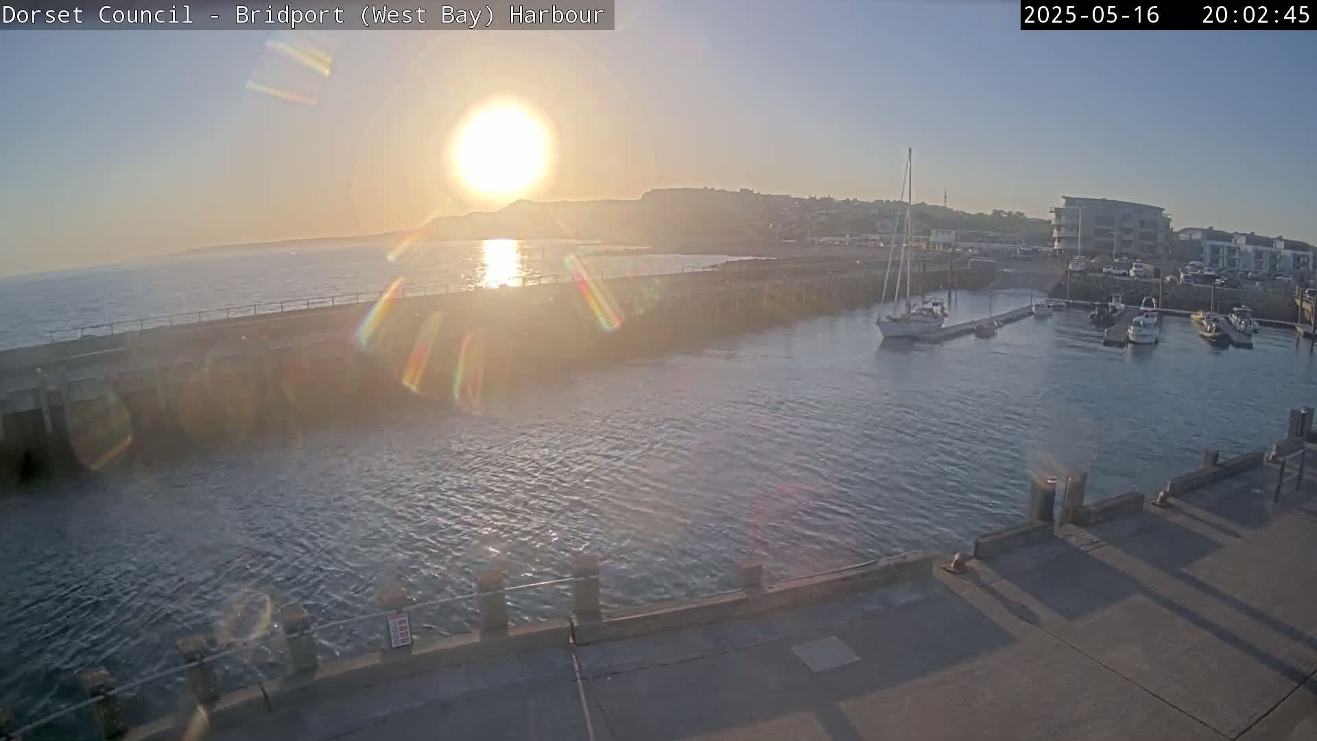 A harbor with several boats is seen at sunset, the sun creating lens flare in the image.