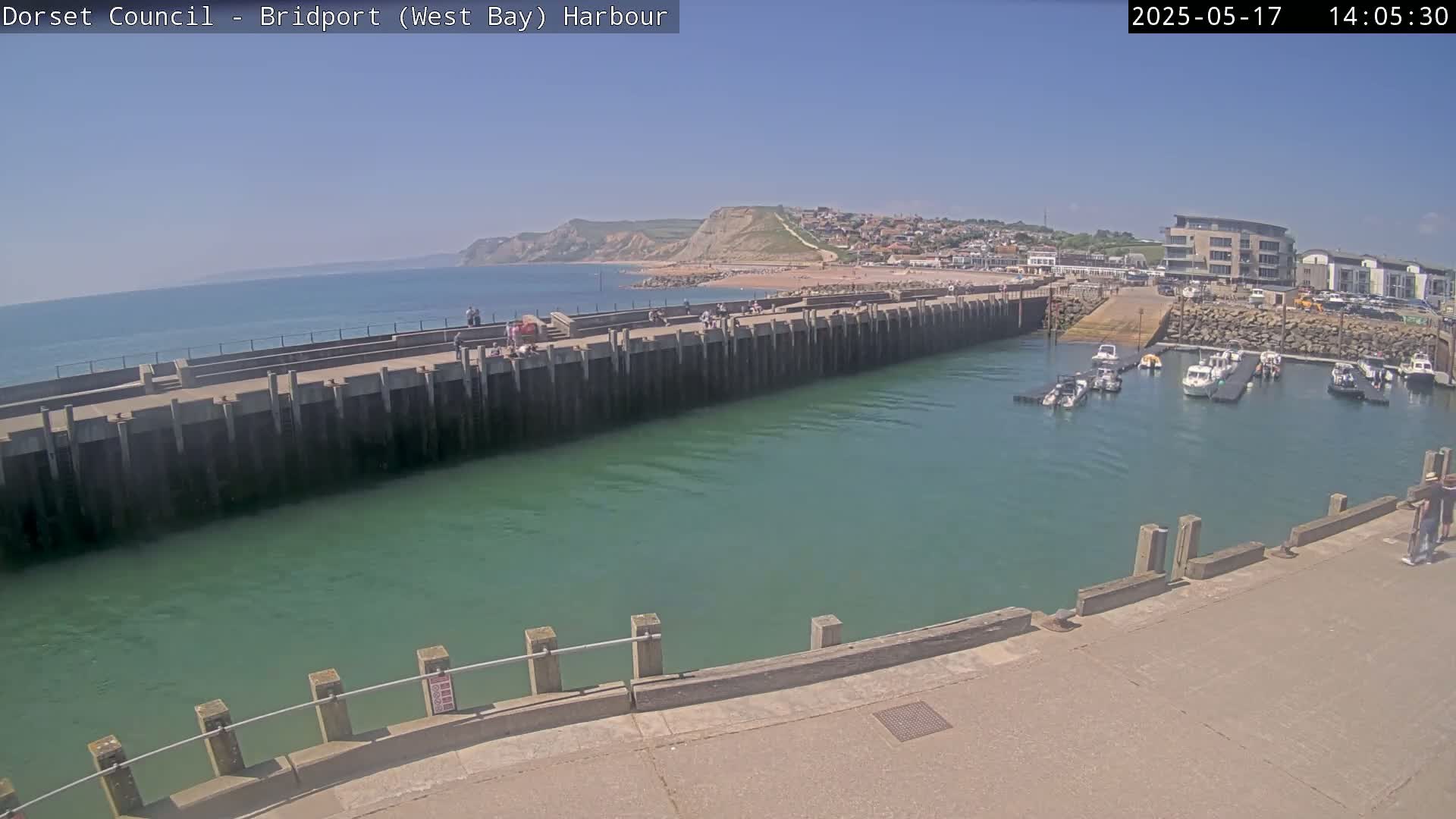 A harbor with calm, green water, several boats docked, a long concrete pier with people on it, and a town and cliffs visible in the distance under a clear, sunny sky.