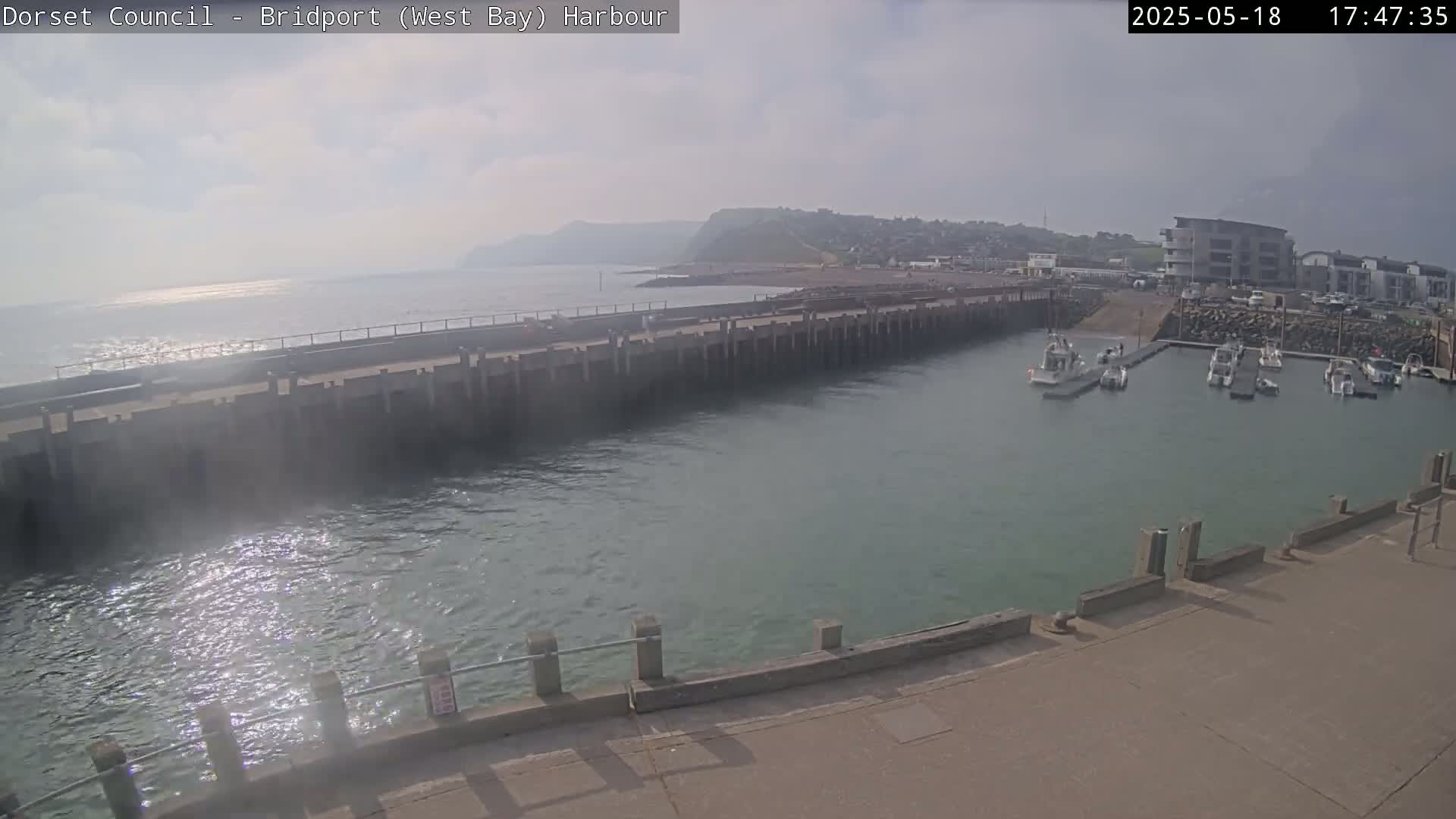 A harbor with several small boats moored, a long concrete pier, and a hazy, overcast sky.