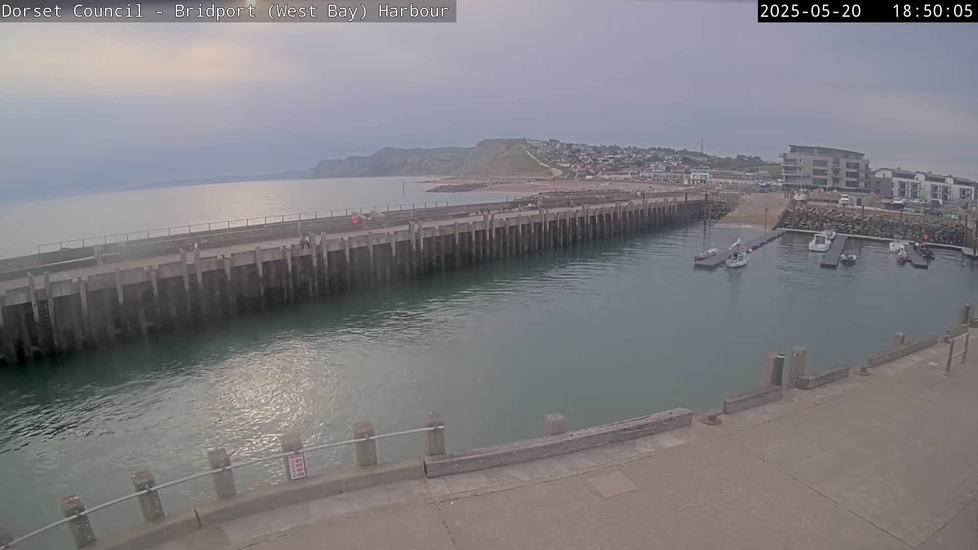 A harbor with several small boats moored, a long concrete pier, and a calm sea under an overcast sky.