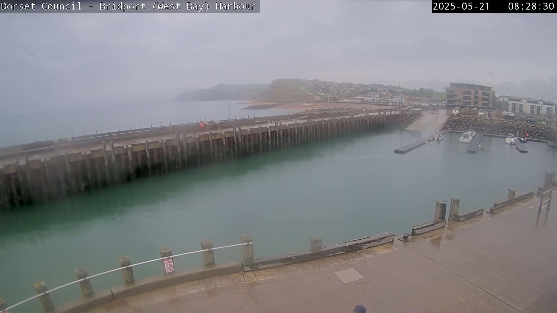 A harbor with a long pier and several boats is visible in overcast, rainy conditions, with a hillside visible in the distance.