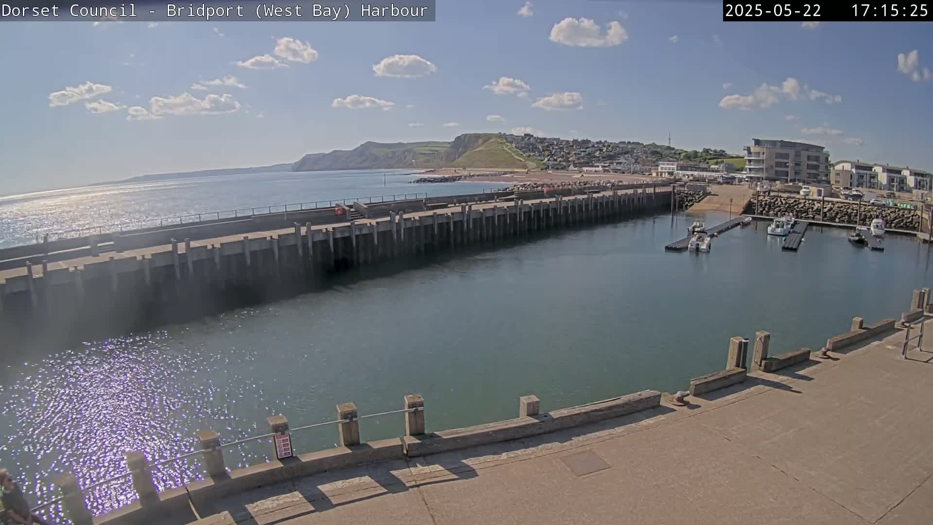 A harbor with calm, sparkling water under a partly sunny sky, featuring a long concrete pier, docked boats, and a town nestled against a hillside in the background.