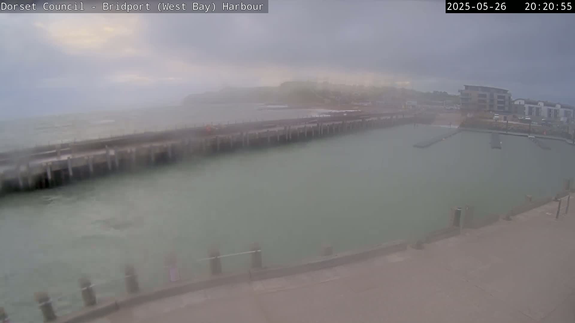 A harbor with a long pier extends into a hazy, overcast sea; buildings are visible in the background.