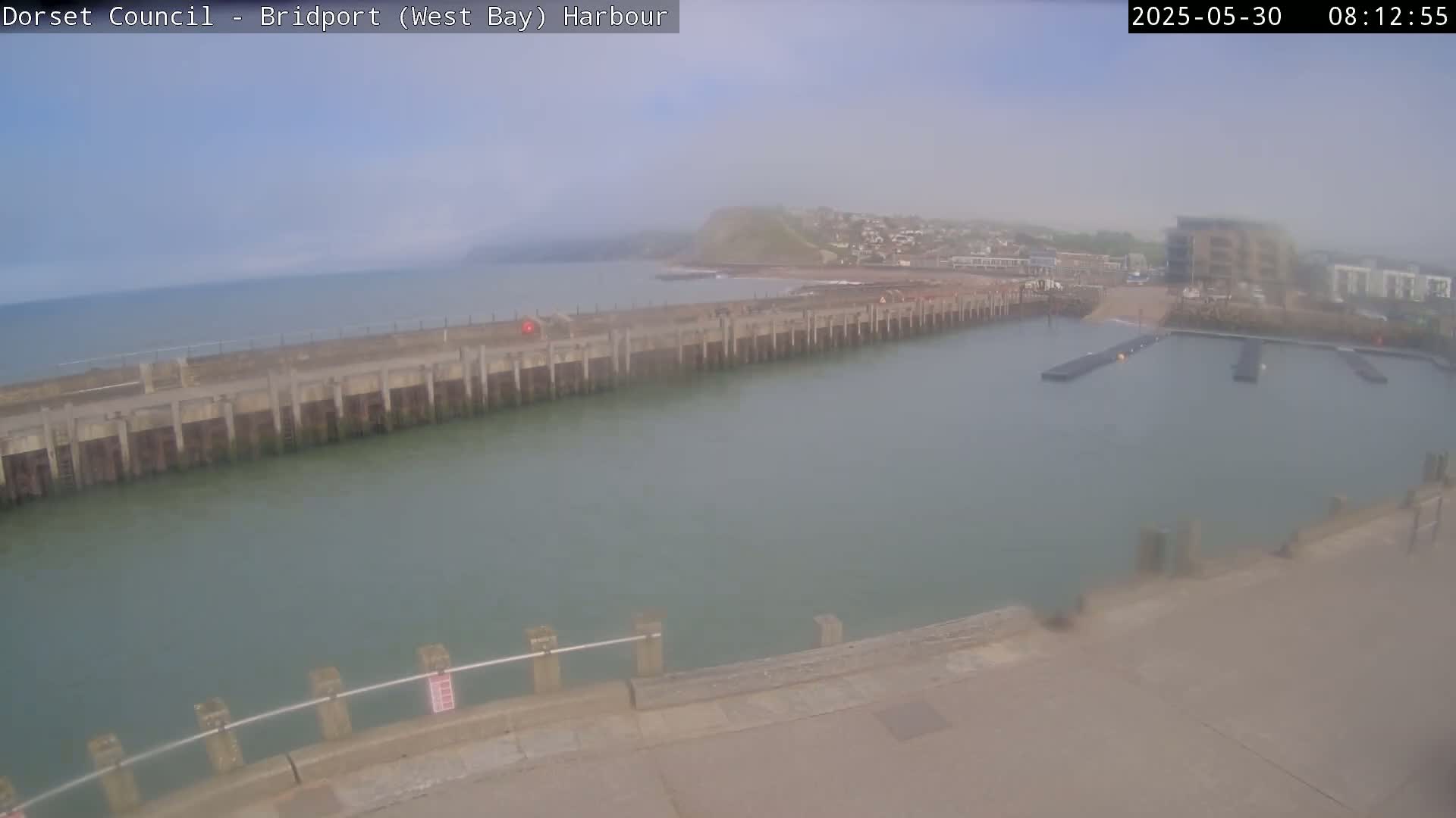 A hazy view shows a harbor with a long pier extending into calm, green-tinged water, a small town and a cliff visible in the background.