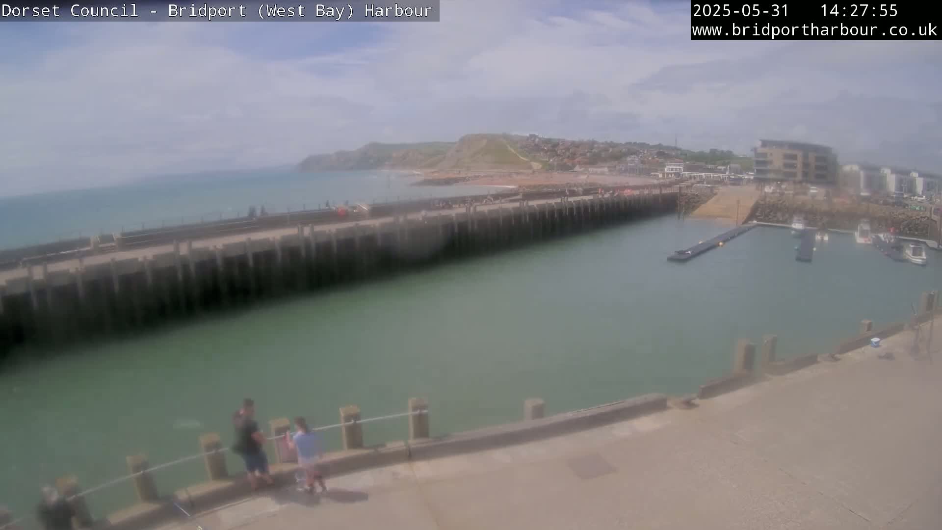 A harbor with calm, green water is seen on a sunny day, with a few people standing near a wall overlooking boats docked in the harbor and a beach in the distance.