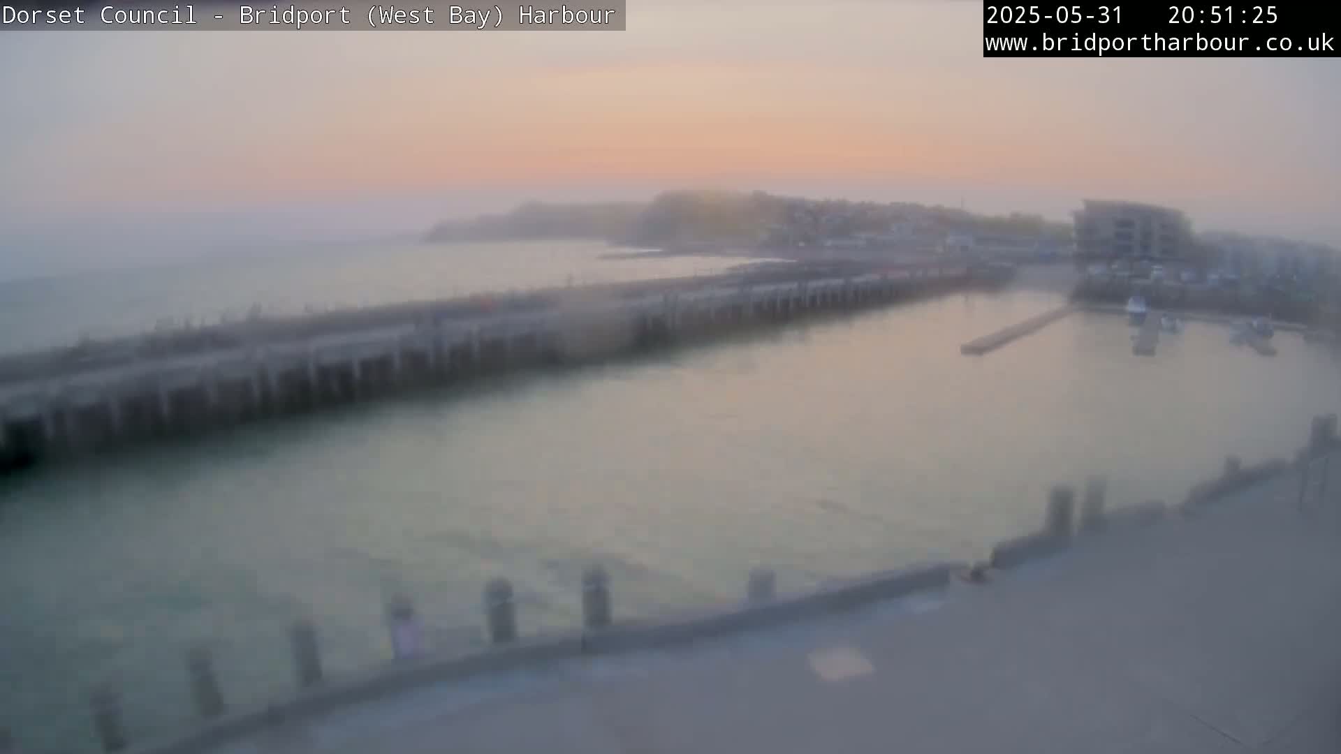 A hazy sunset illuminates a harbor with calm water, a long pier, and a few boats.