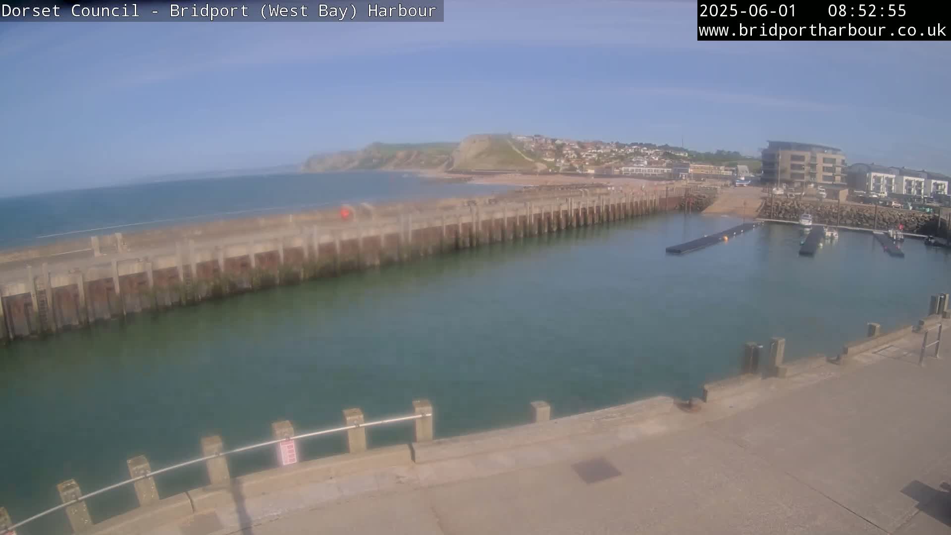A harbor with calm, green water is seen next to a sandy beach and a town under a clear, sunny sky.