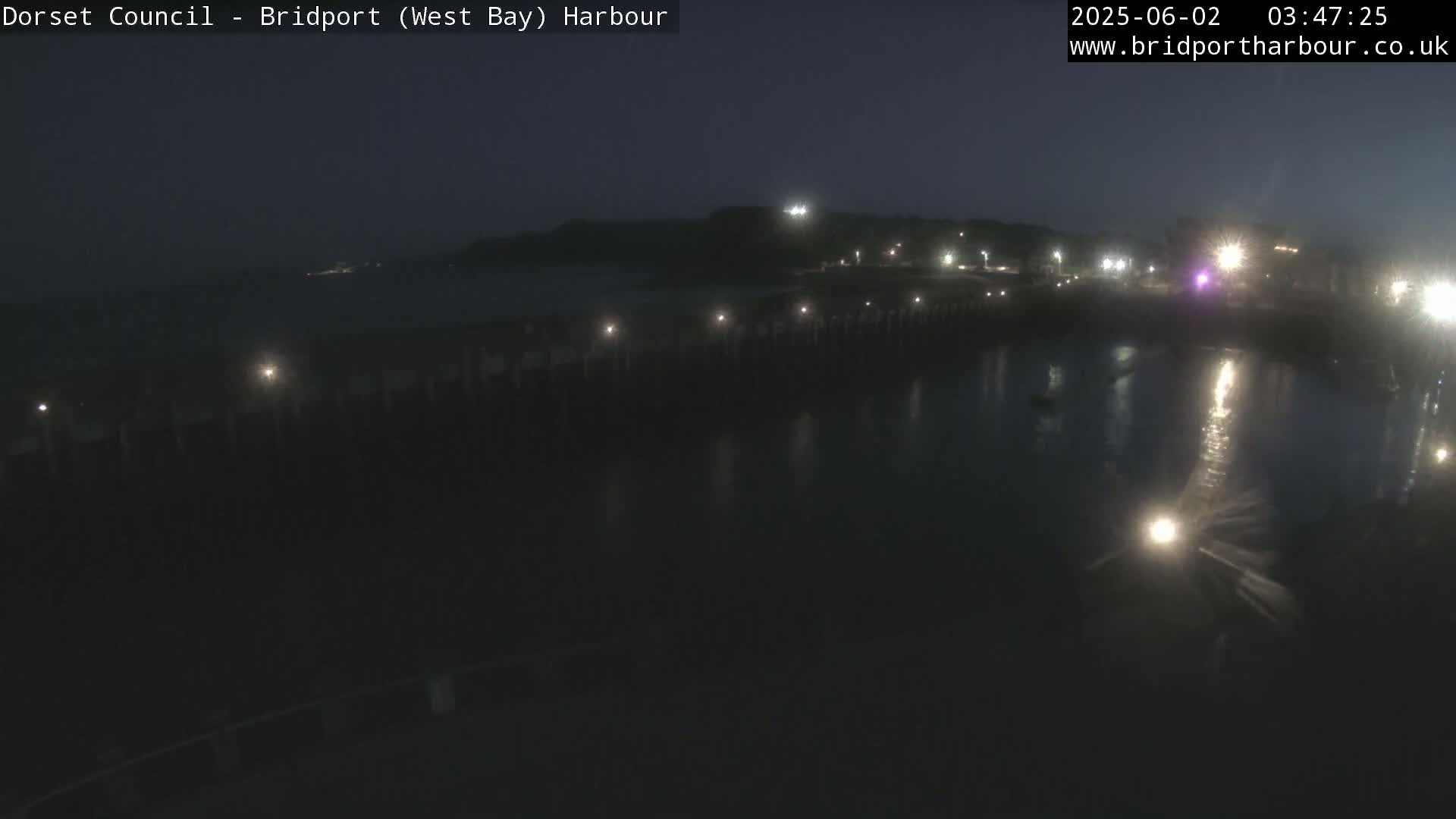 A dark nighttime harbor scene shows a long pier with lights, a calm body of water reflecting the lights, and a hill in the background under a clear, dark sky.