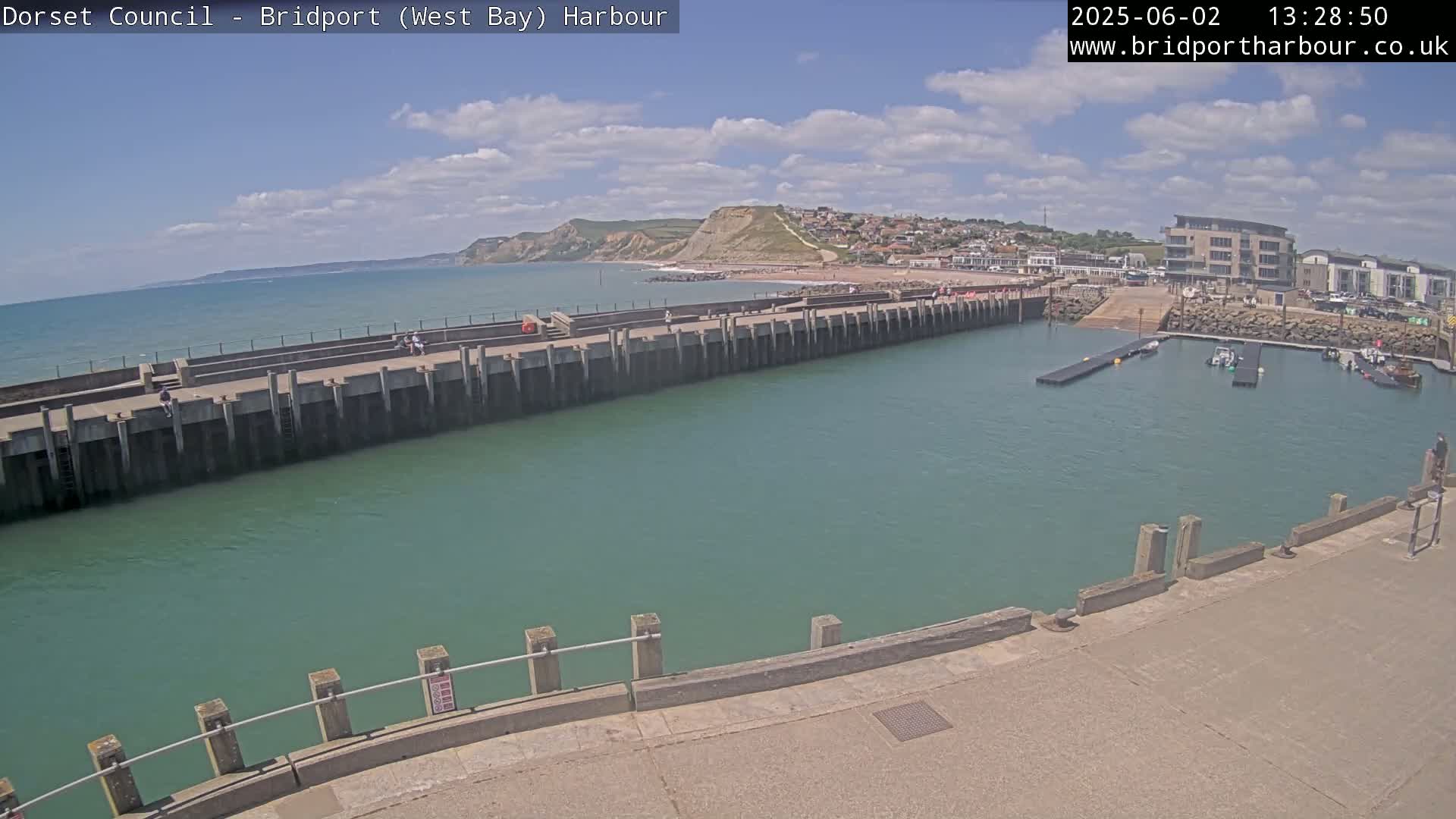 A harbor with a calm, teal-green waterway, a long concrete pier, and a view of a beach and cliffs under a partly cloudy sky.