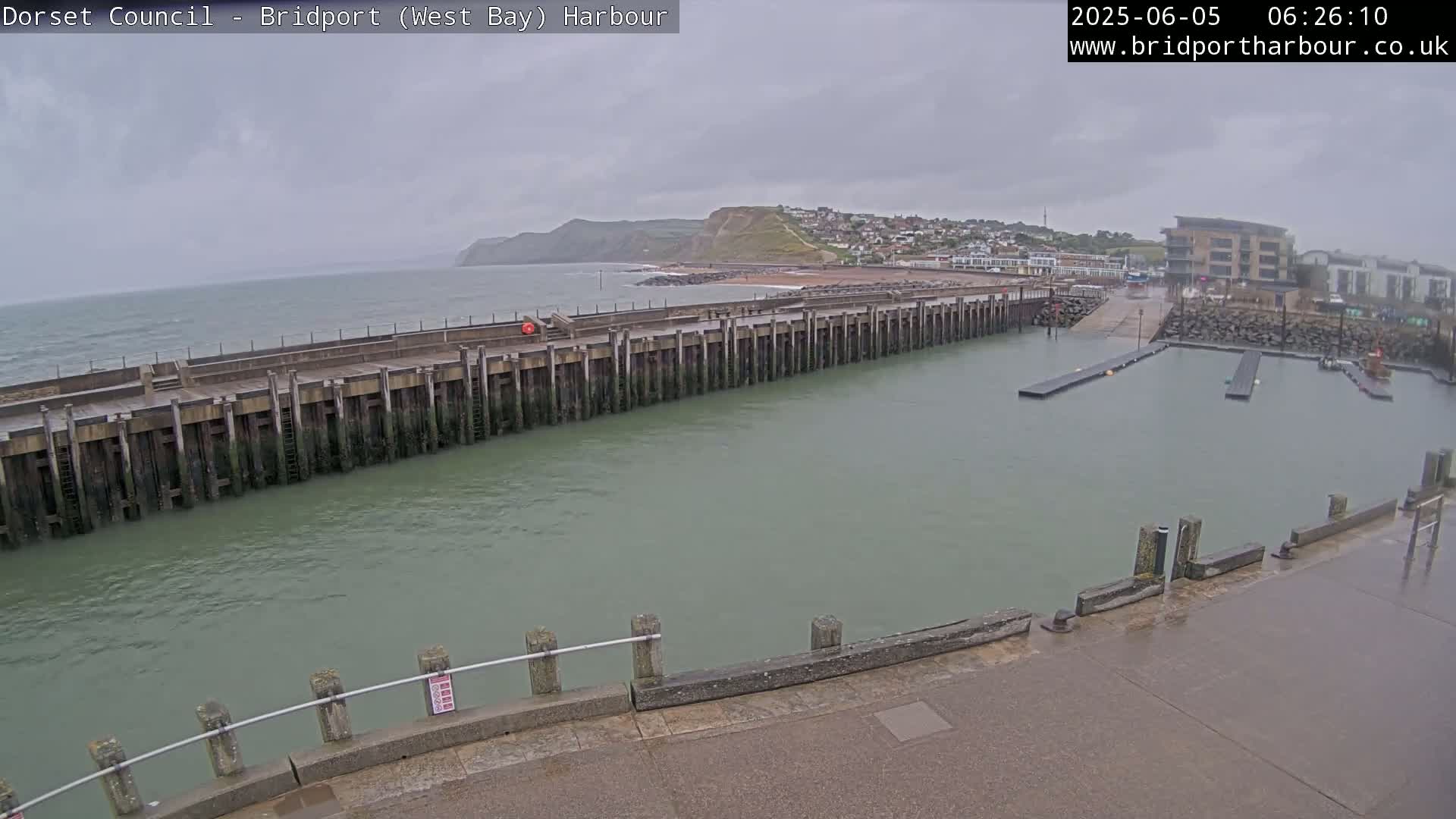 A harbor with a long pier extending into calm, greenish water under an overcast sky, showing a town and hills in the background.