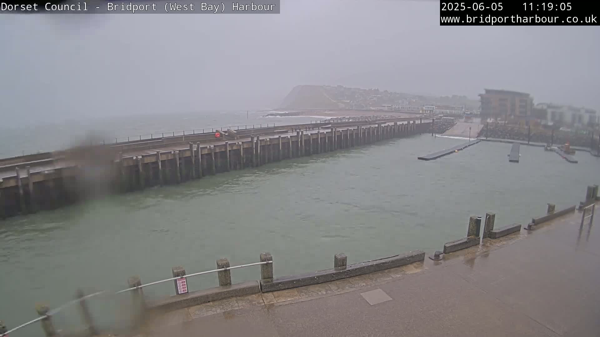 A harbor entrance with a long pier is seen on an overcast, rainy day with choppy water.