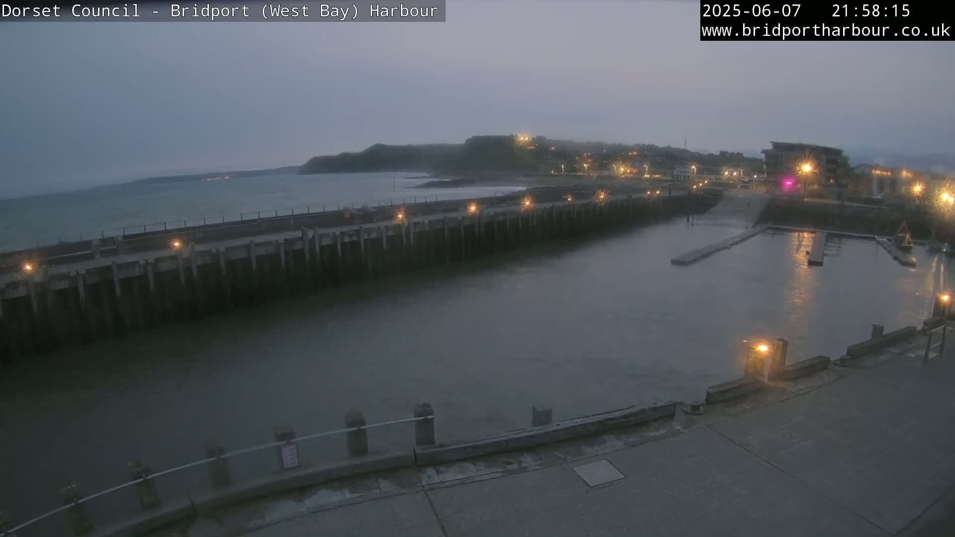 A dimly lit harbor at twilight, with calm water and a dark hill visible in the background.