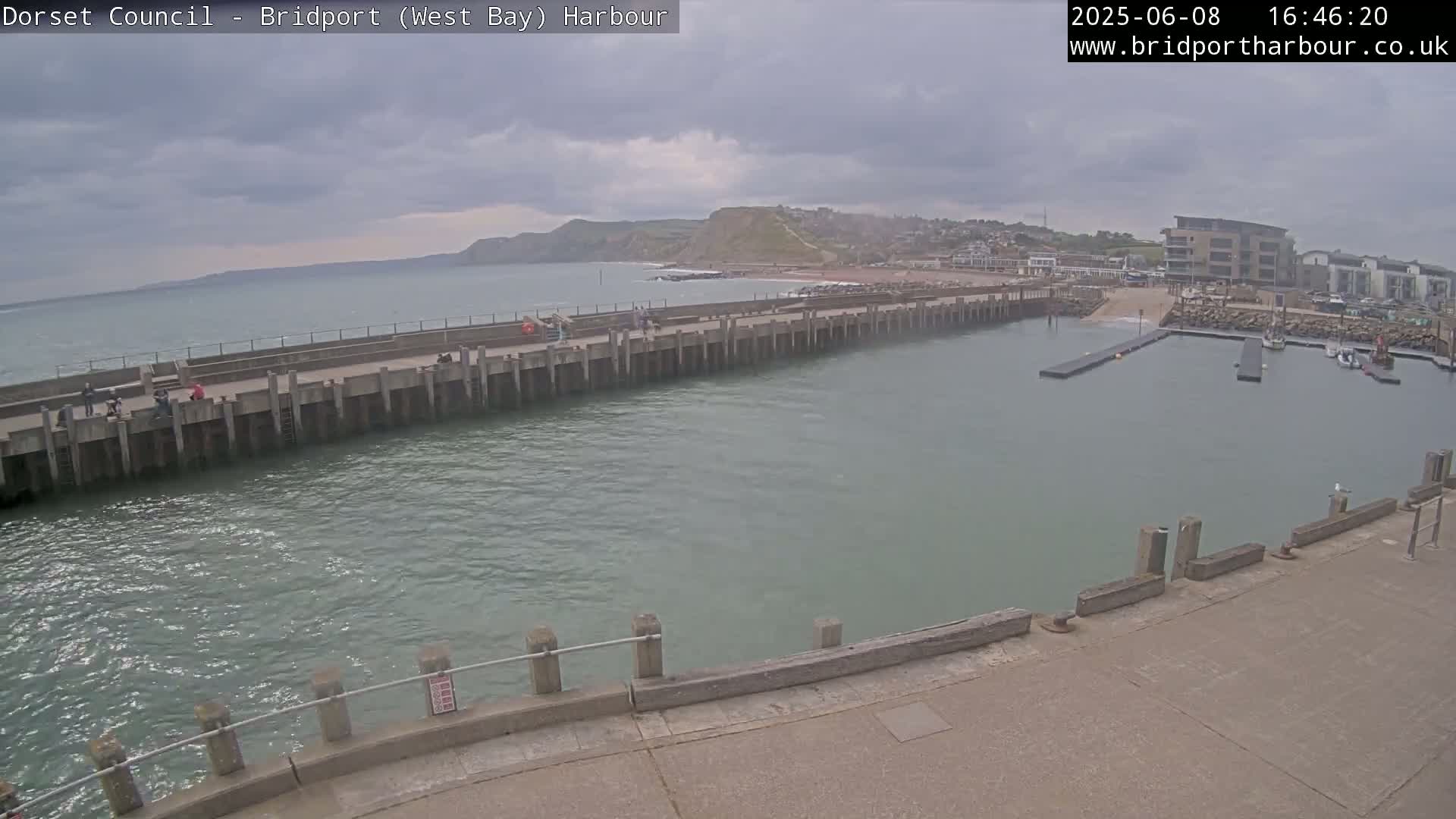 A harbor with a long concrete pier extends into calm, greenish-gray water under an overcast sky, with a small town and beach visible in the background.