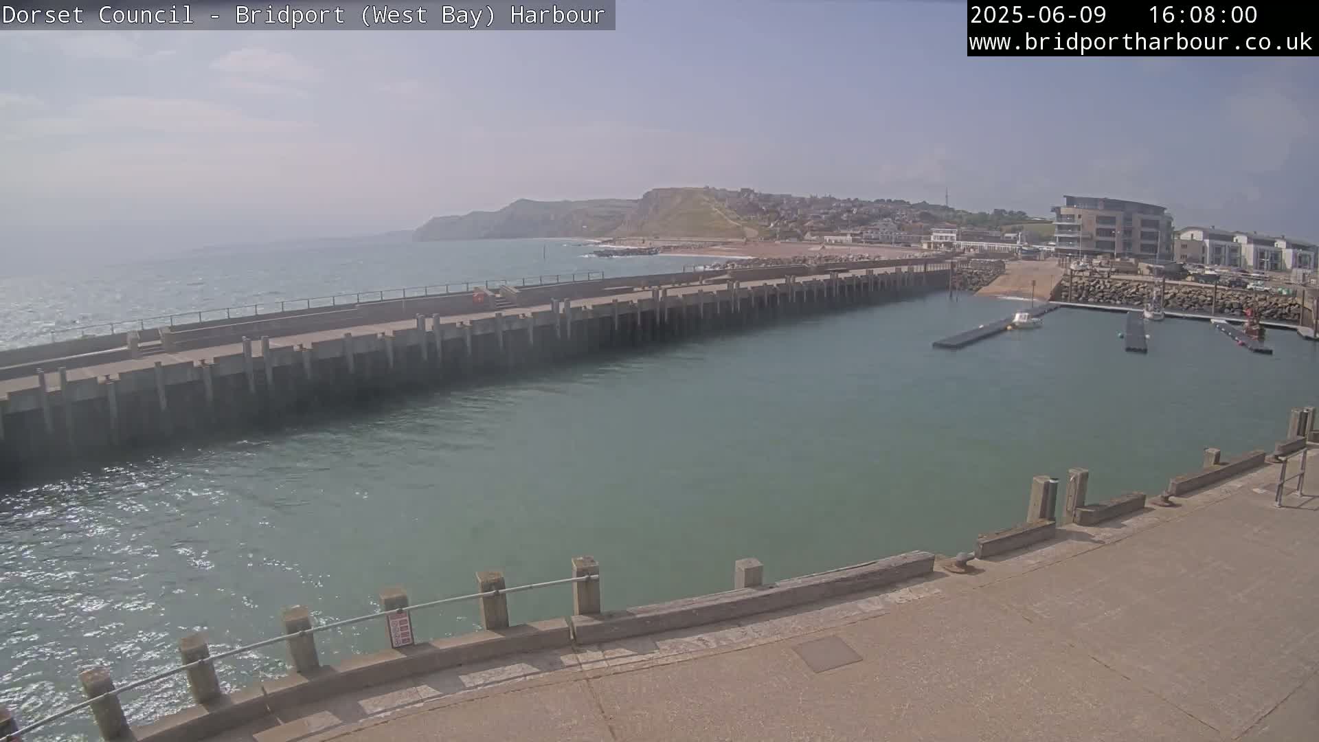 A harbor with a long concrete pier juts into calm, greenish-blue water under a mostly sunny sky, with a town and hills visible in the distance.