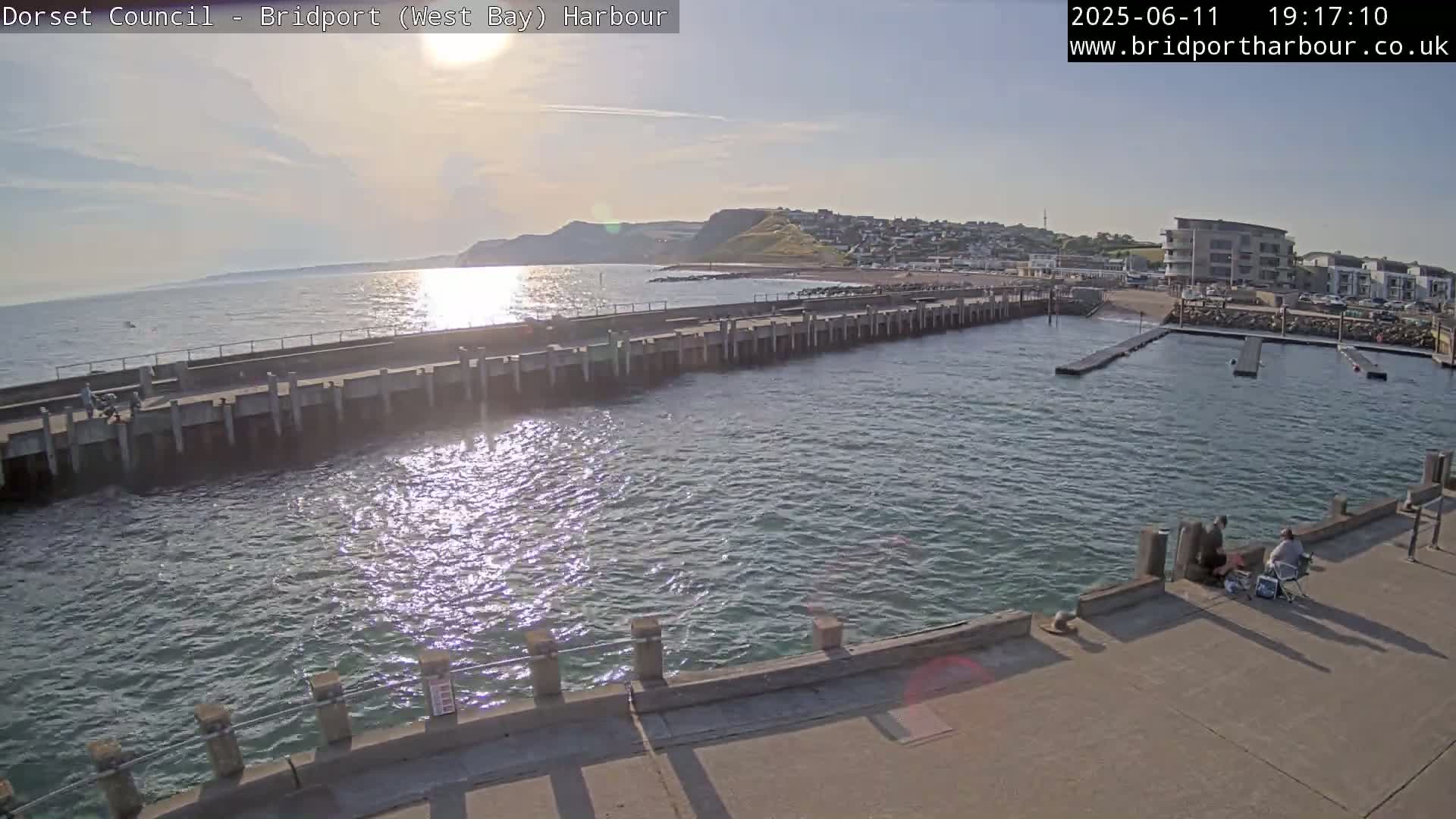 A harbor with calm, sunlit water is shown, featuring a long pier extending into the water, a coastal town in the background, and two people sitting on the pier.