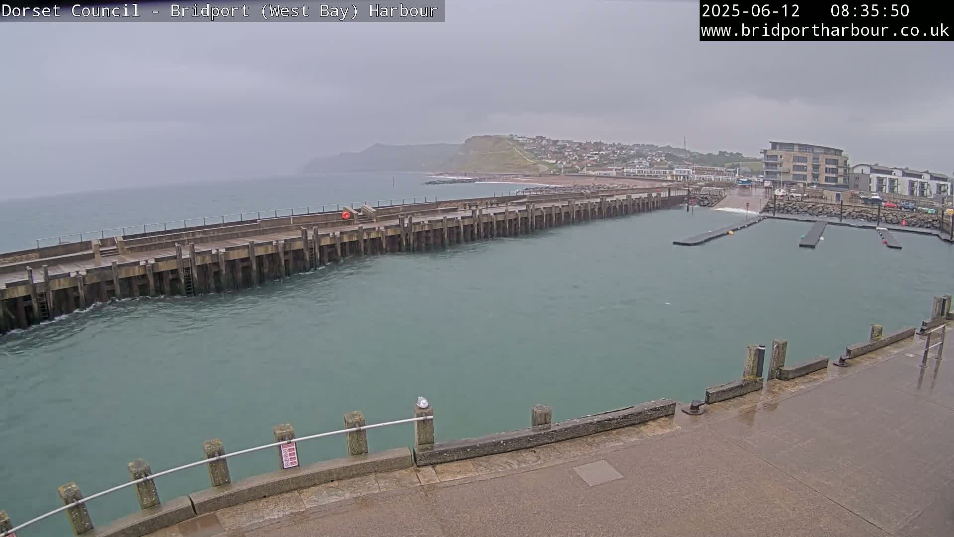 A harbor with a long pier extending into choppy, greenish-grey water under an overcast sky.