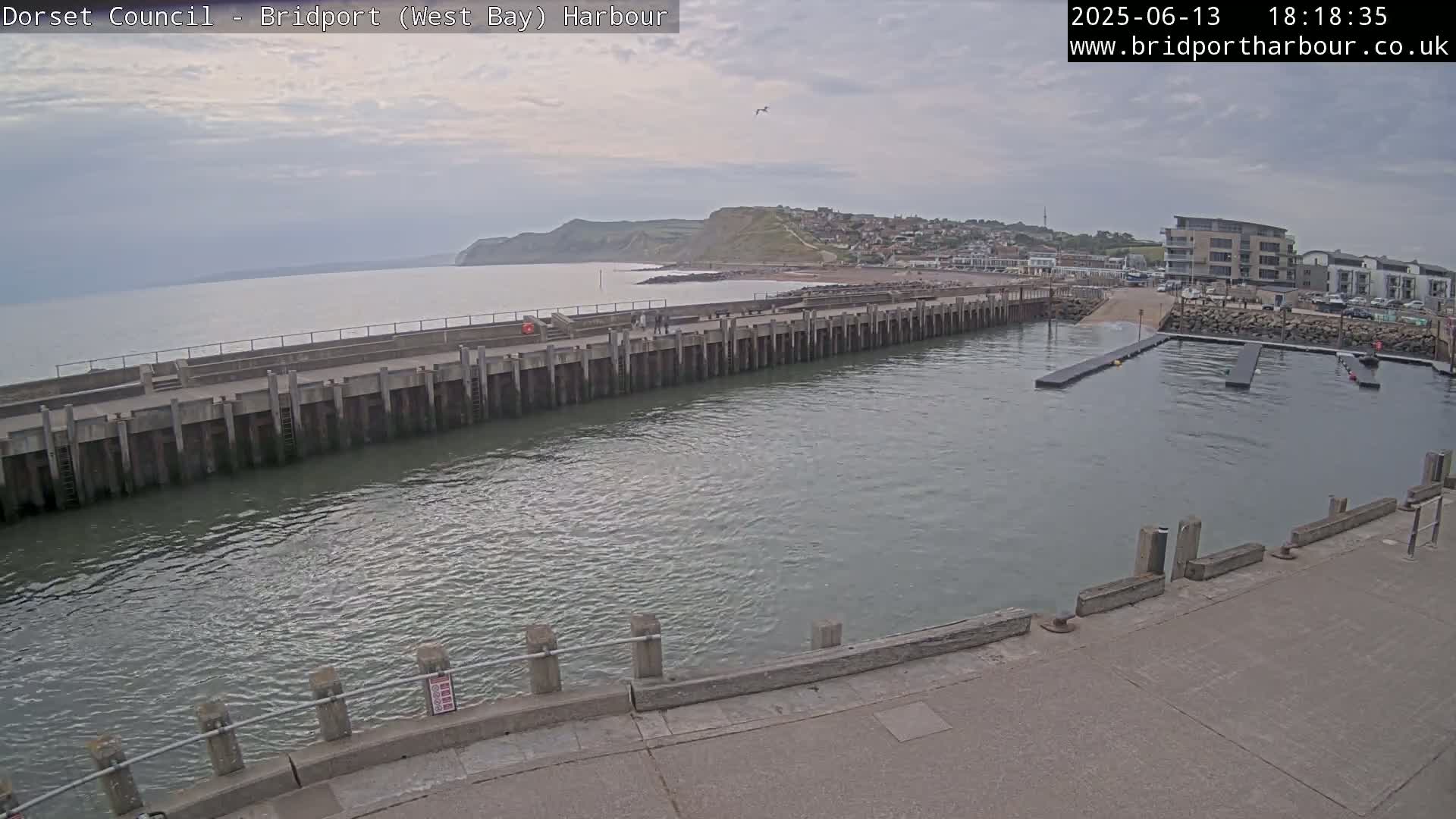 A harbor with a long concrete wall, calm water, and a distant coastline under an overcast sky.