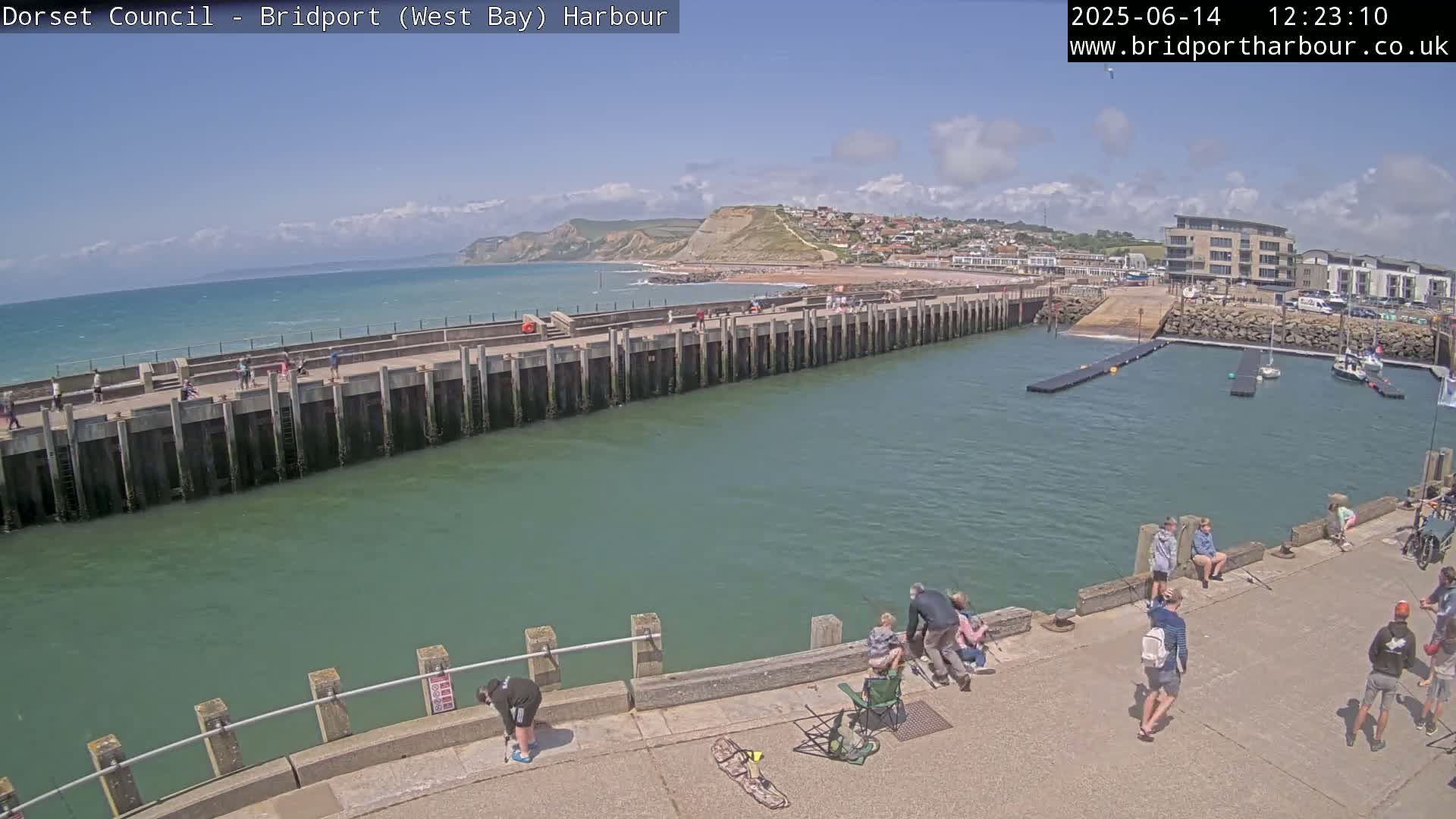 A harbor with a long concrete pier, people fishing and walking along the pier, and boats in the marina, all under a mostly sunny sky.