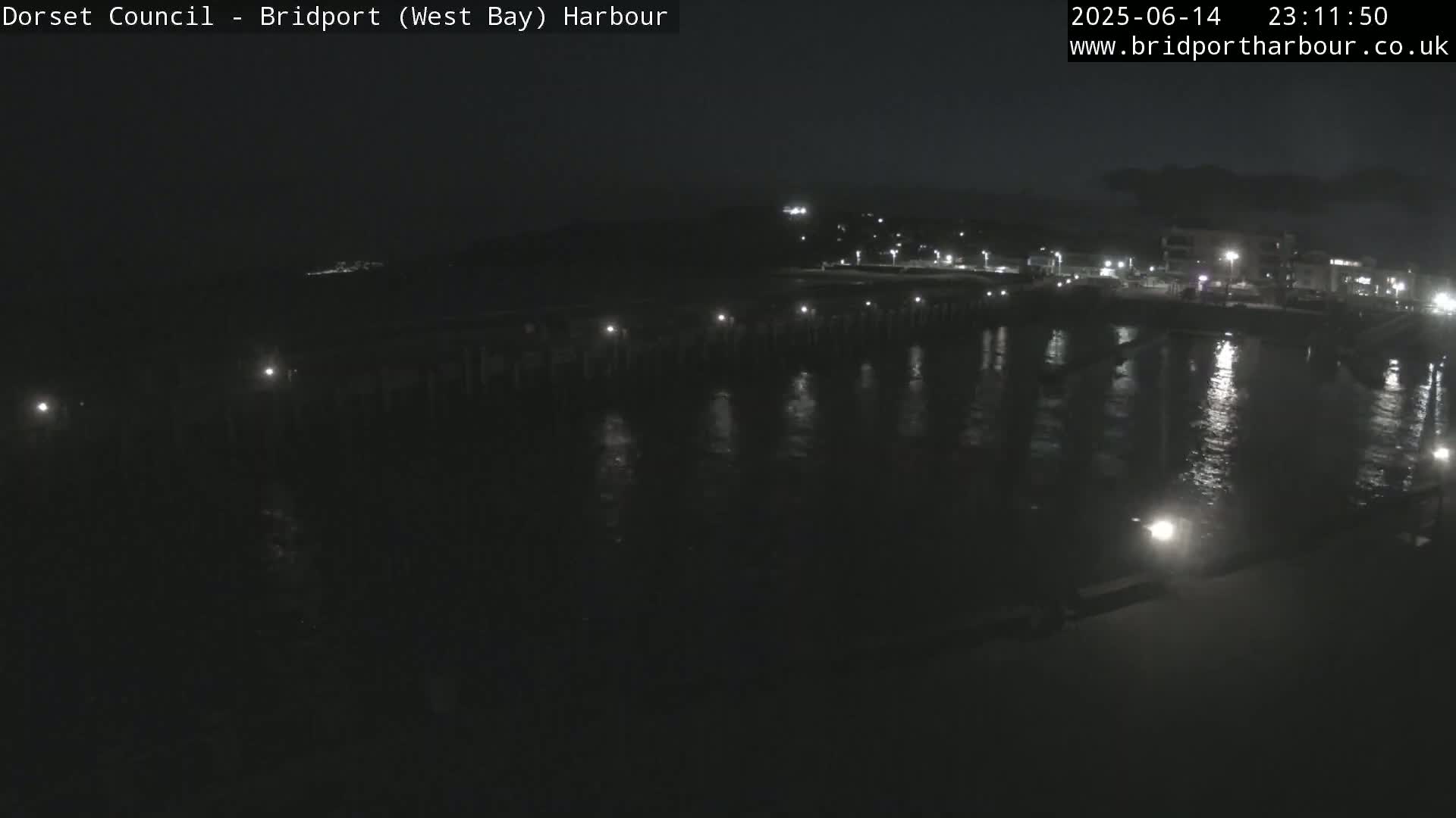 A harbor with a long pier, calm water, and a partly sunny sky overlooks a beach and coastal town.