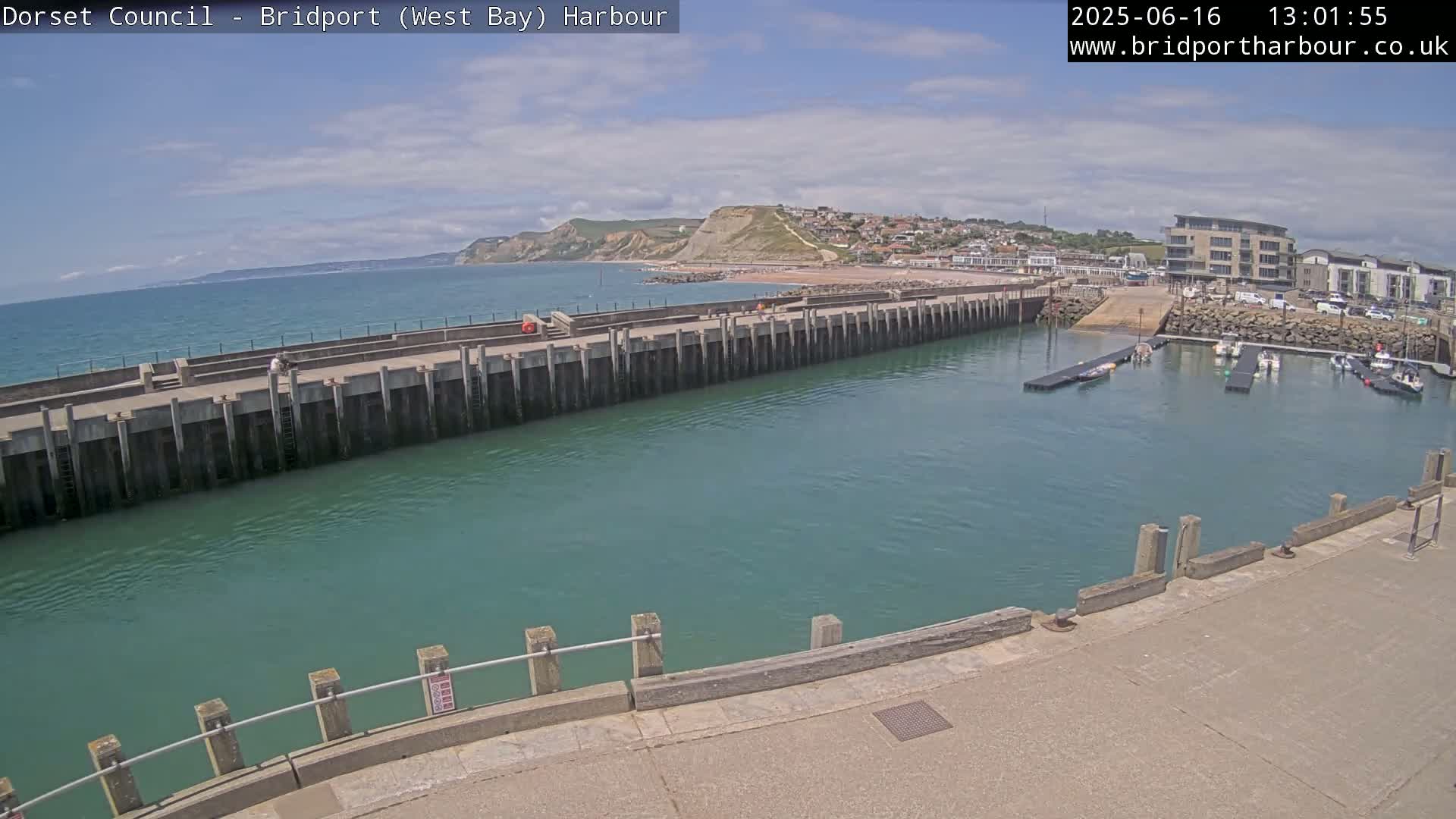 A harbor with a long concrete pier extending into teal water, under a mostly sunny sky, showing a small town and cliffs in the background.