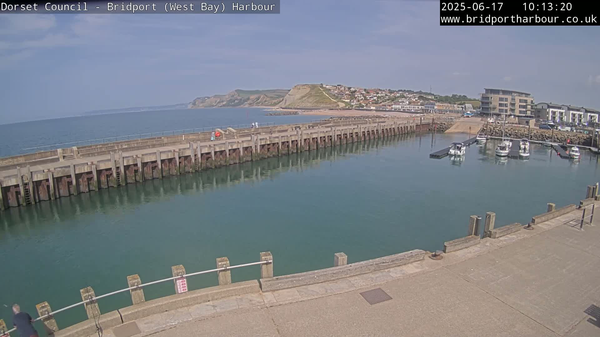 A harbor with calm, teal water, a long concrete pier, several boats docked in the harbor, and a coastal town visible in the background on a sunny day.