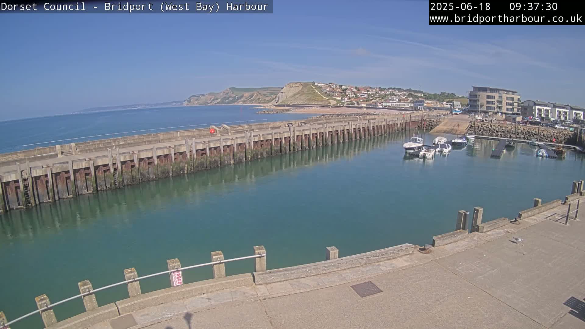 A harbor with several boats is shown on a sunny day, with a long concrete wall separating the harbor from the ocean and a coastal town visible in the background.