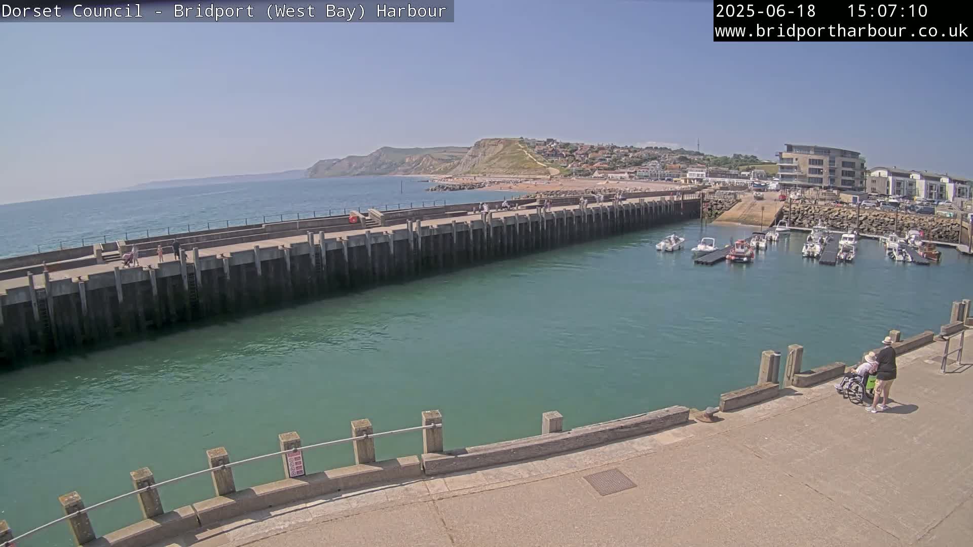 A harbor with several boats docked, a long concrete wall separating the harbor from the ocean, and a person pushing someone in a wheelchair along a walkway on a sunny day.