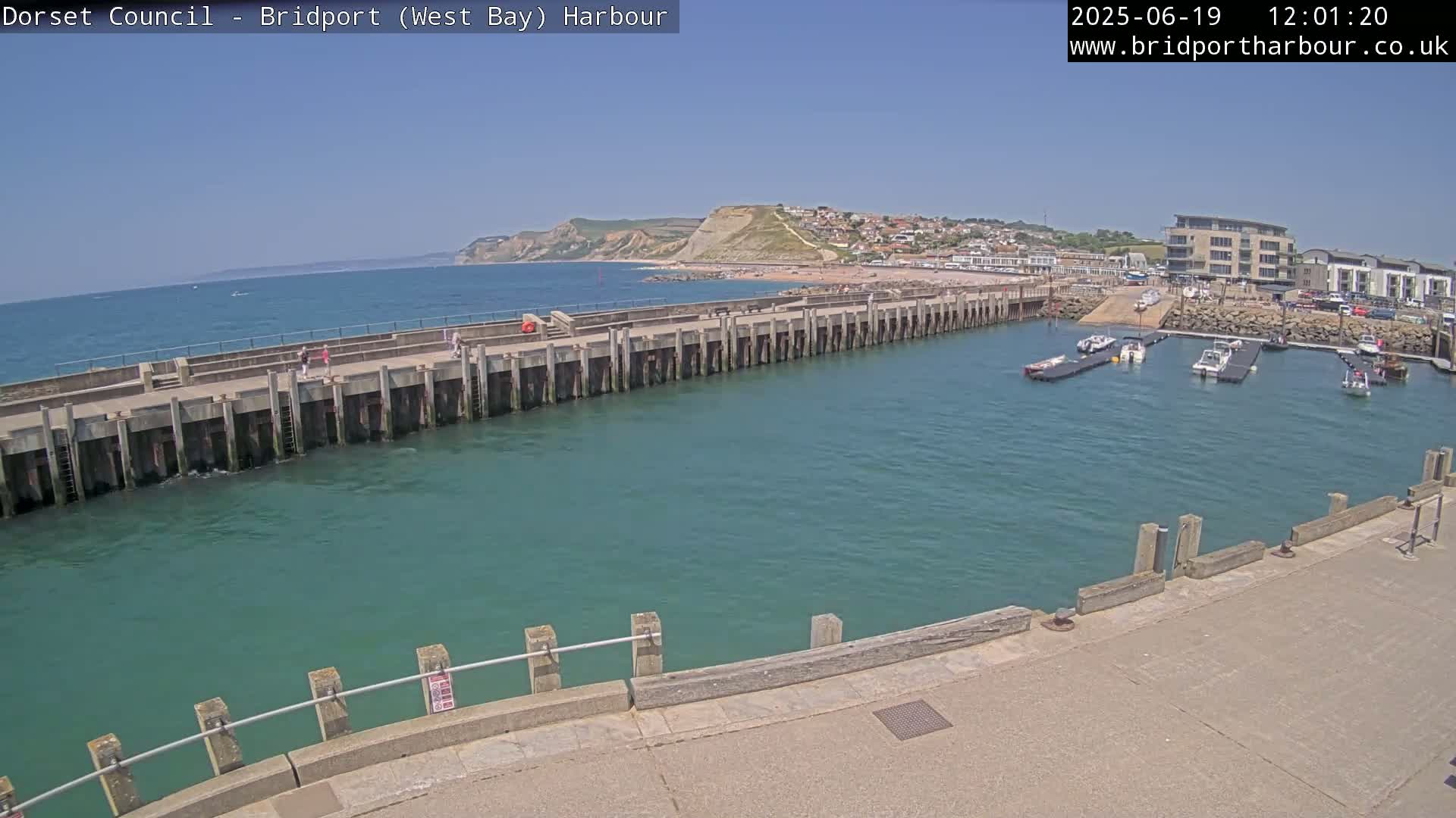 A harbor with calm, teal water is partially enclosed by a long concrete pier under a clear blue sky, with several boats moored in the harbor and a beach visible beyond it.