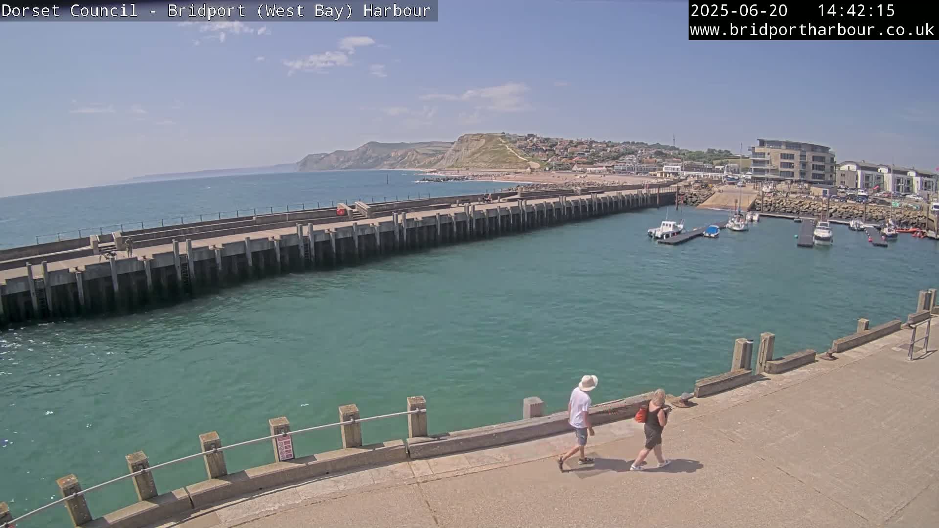 A harbor with calm, teal water, a long concrete pier, several boats moored in the harbor, a sandy beach and cliffs in the distance under a sunny sky.