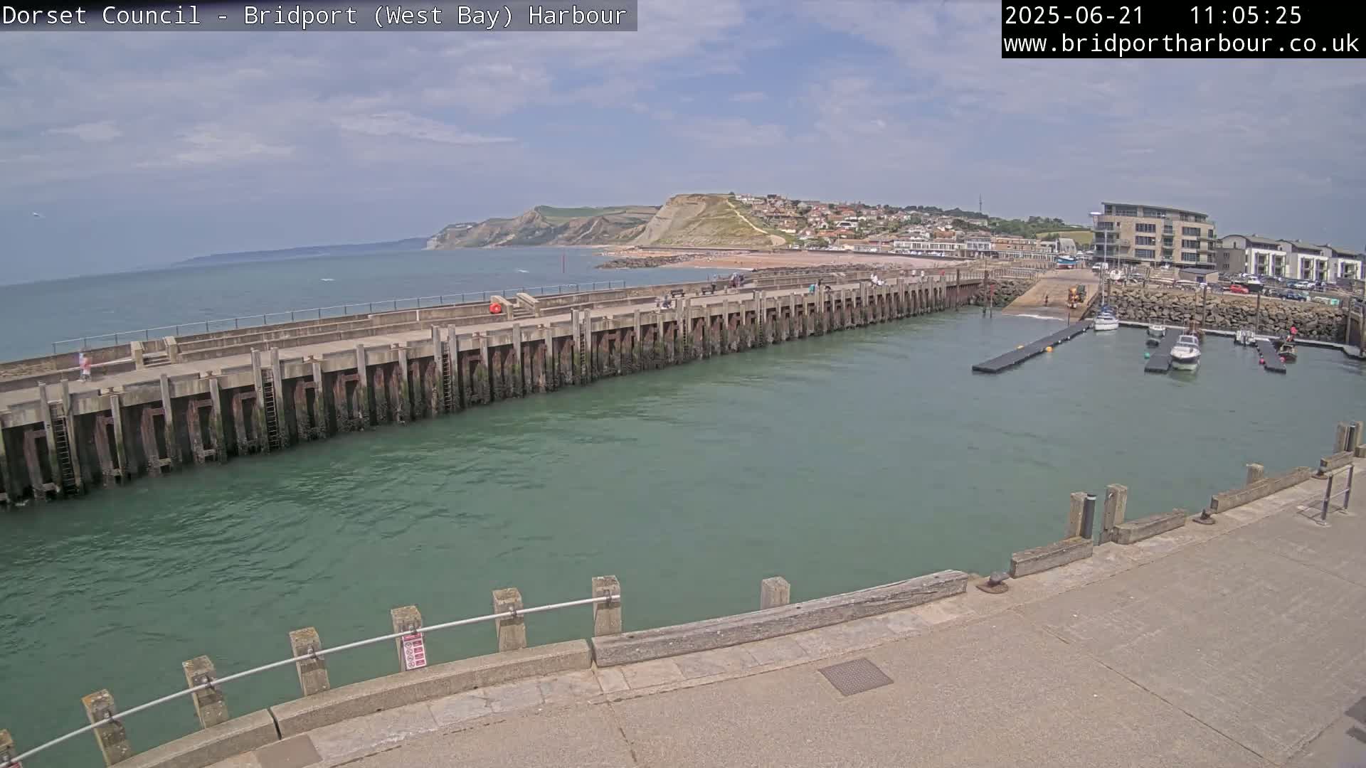 A harbor with a long pier, a calm sea, and a town nestled against a cliff under a partly cloudy sky.